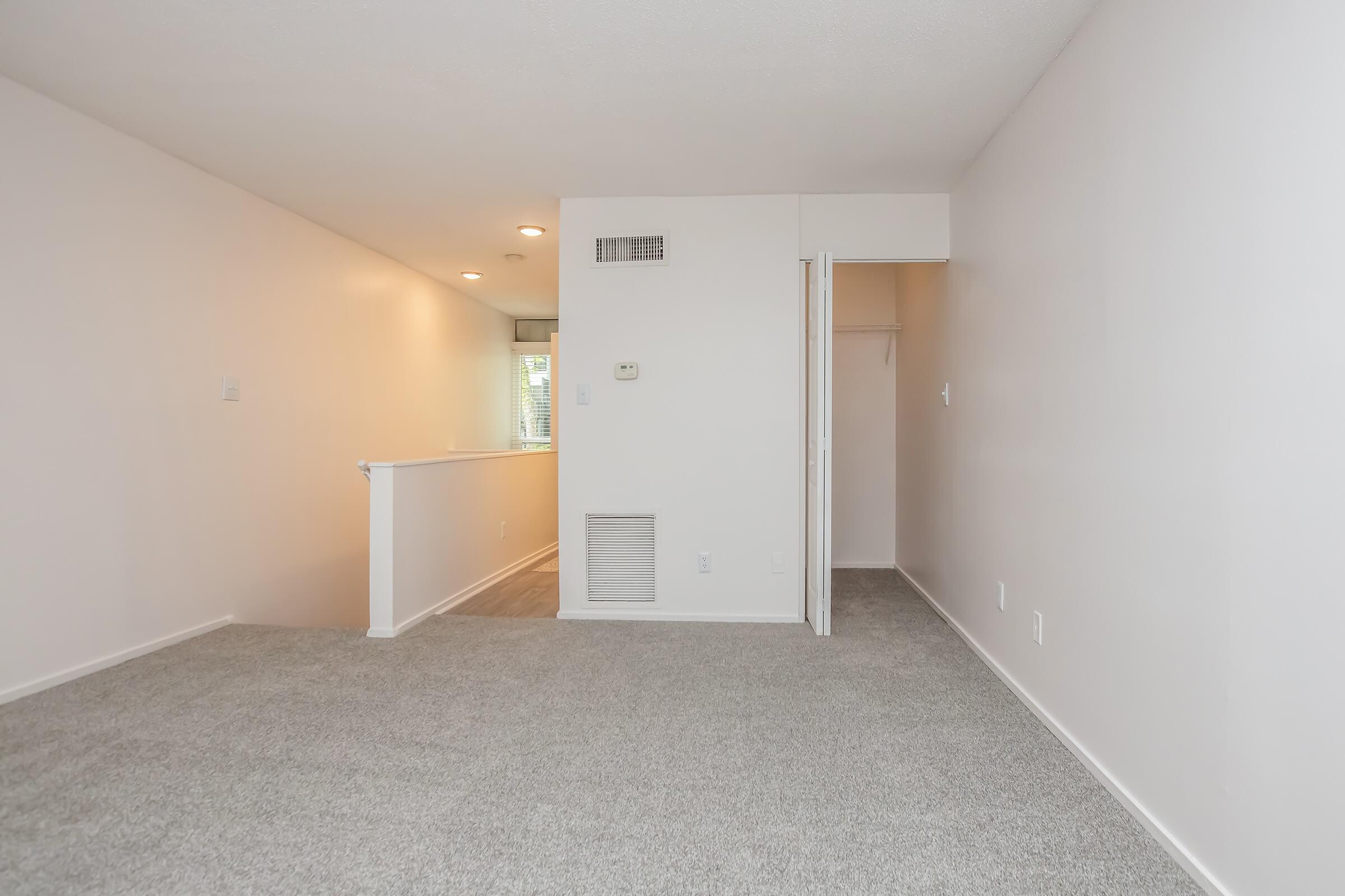 A spacious, well-lit living area with neutral-colored walls and carpet. There's a small entryway leading to a closet and an air vent. Natural light streams in from a window in the background, creating a warm atmosphere. The space is tidy and unfurnished, ready for personal decoration.