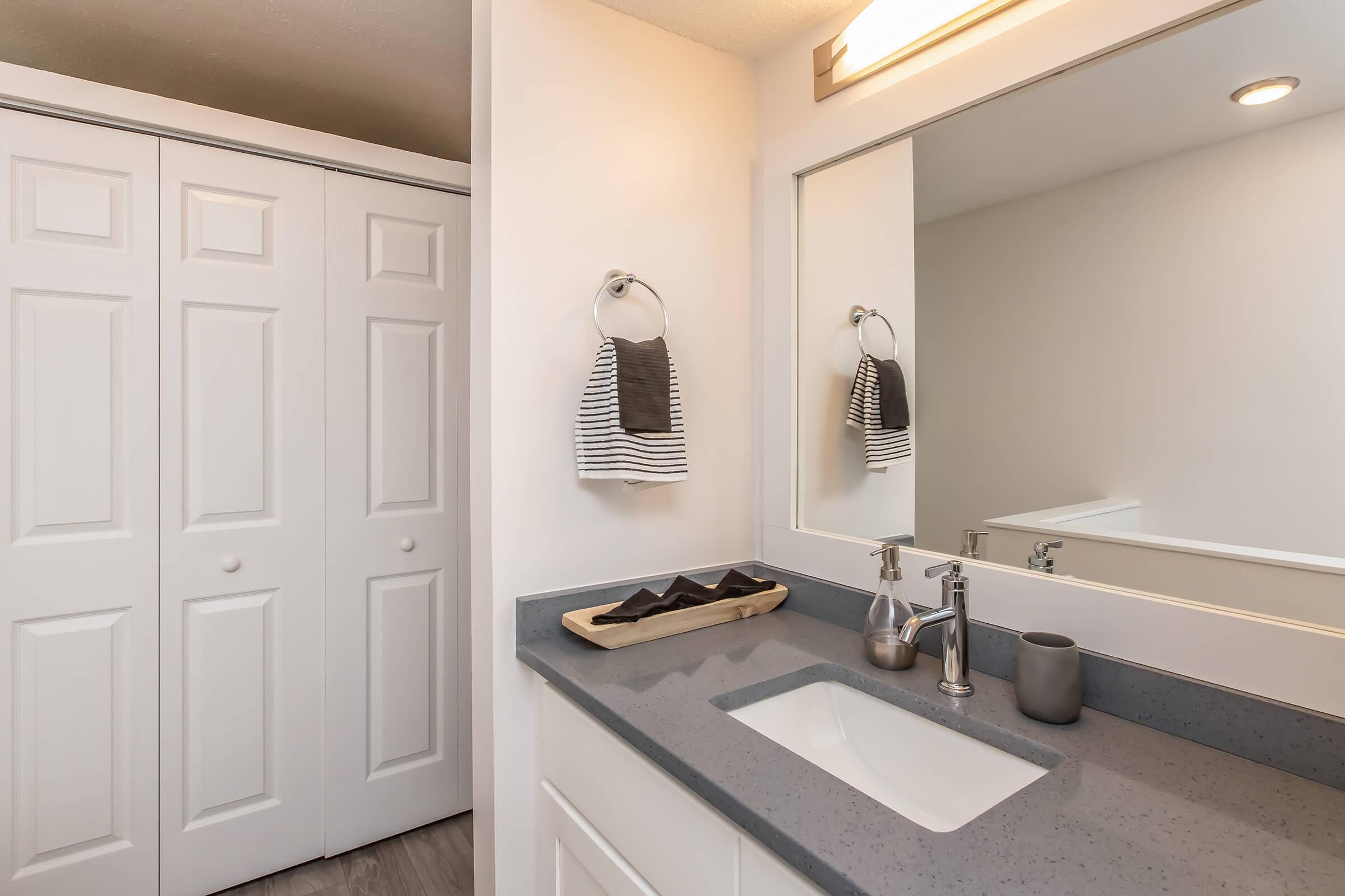 A modern bathroom featuring a minimalist design. It has a gray countertop with a sink, a towel rack with striped towels, and a neatly organized decorative tray. The wall is painted white, and there are double closet doors in the background. A mirror above the sink enhances the bright ambiance.
