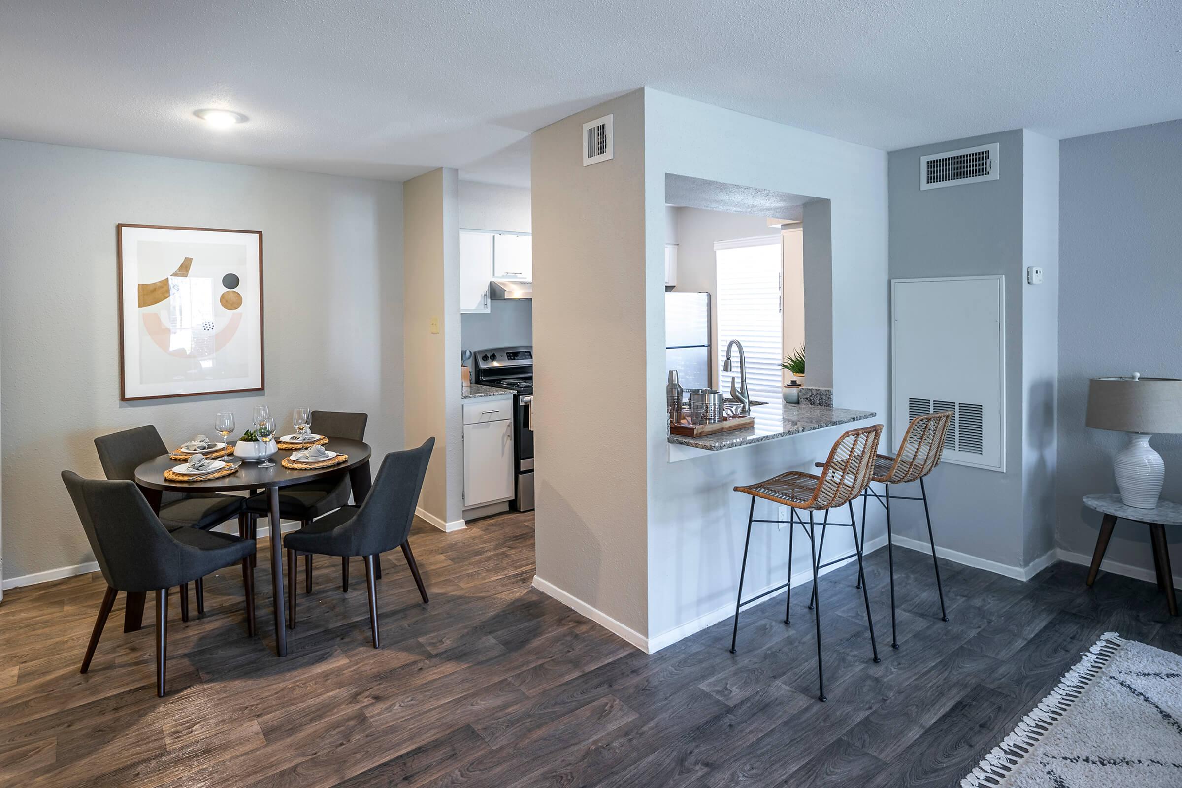 A modern dining area with a round table set for four, featuring plates and glasses. Adjacent is a kitchen with white cabinetry and stainless steel appliances. A breakfast bar with two stools is visible. The room has neutral-toned walls and hardwood-style flooring, creating a welcoming atmosphere.