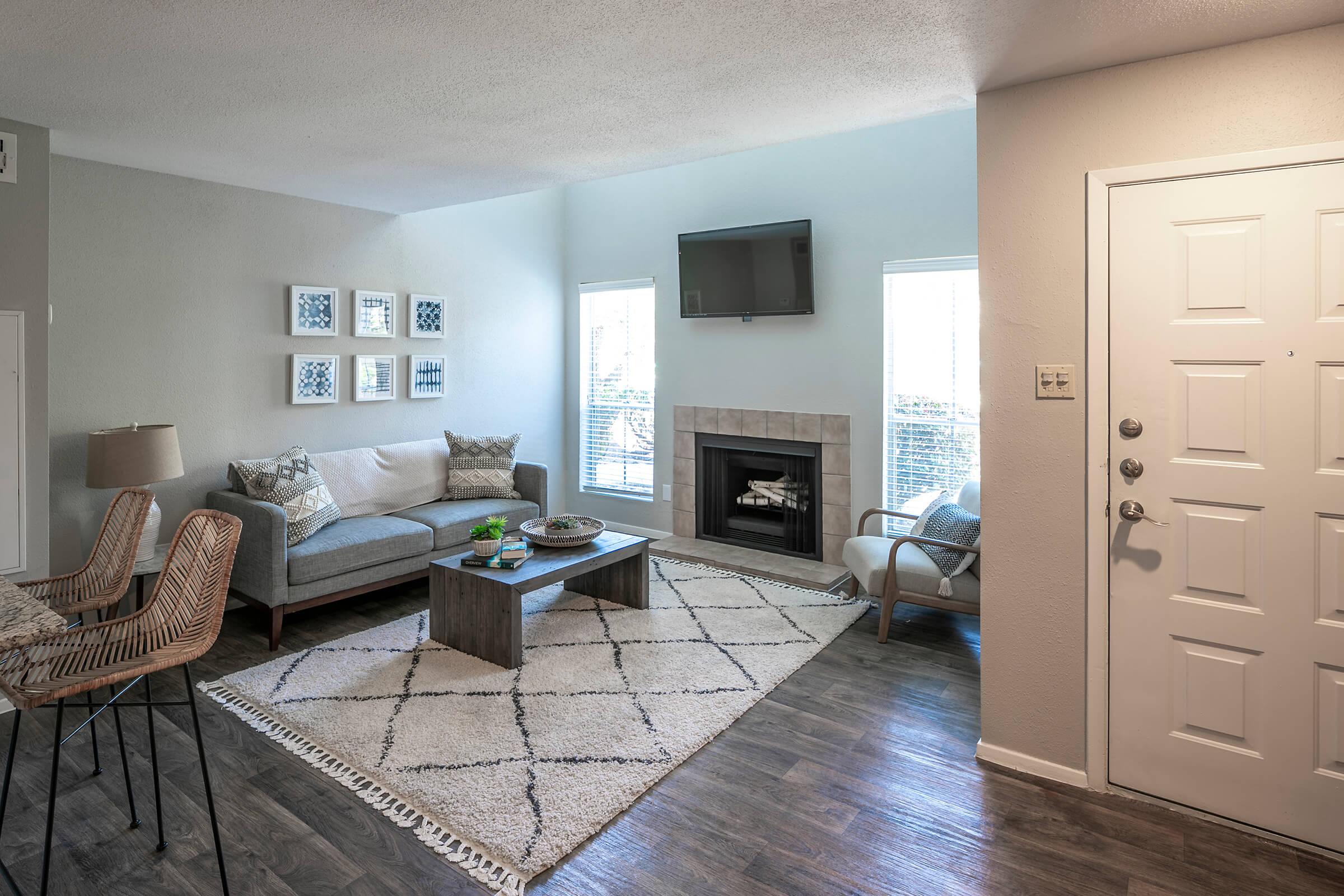 Cozy living room featuring a light gray sectional sofa, a wooden coffee table on a patterned rug, and modern decor including wall art. There is a fireplace beneath a wall-mounted TV, large windows allowing natural light, and a stylish entryway with a door and a small seating area.