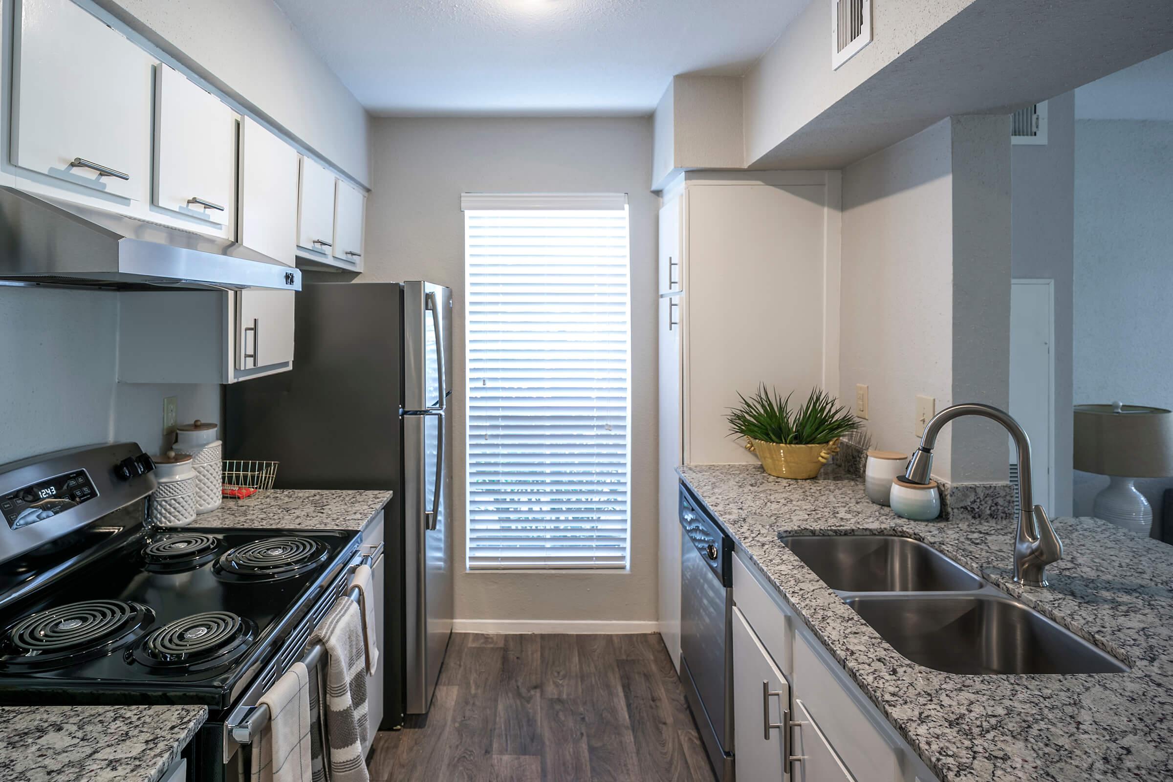 Modern kitchen featuring a black electric stove, stainless steel refrigerator, and double sink with a contemporary faucet. Grey granite countertops complement white cabinetry. A window with horizontal blinds provides natural light, while a small potted plant adds a touch of greenery to the space.