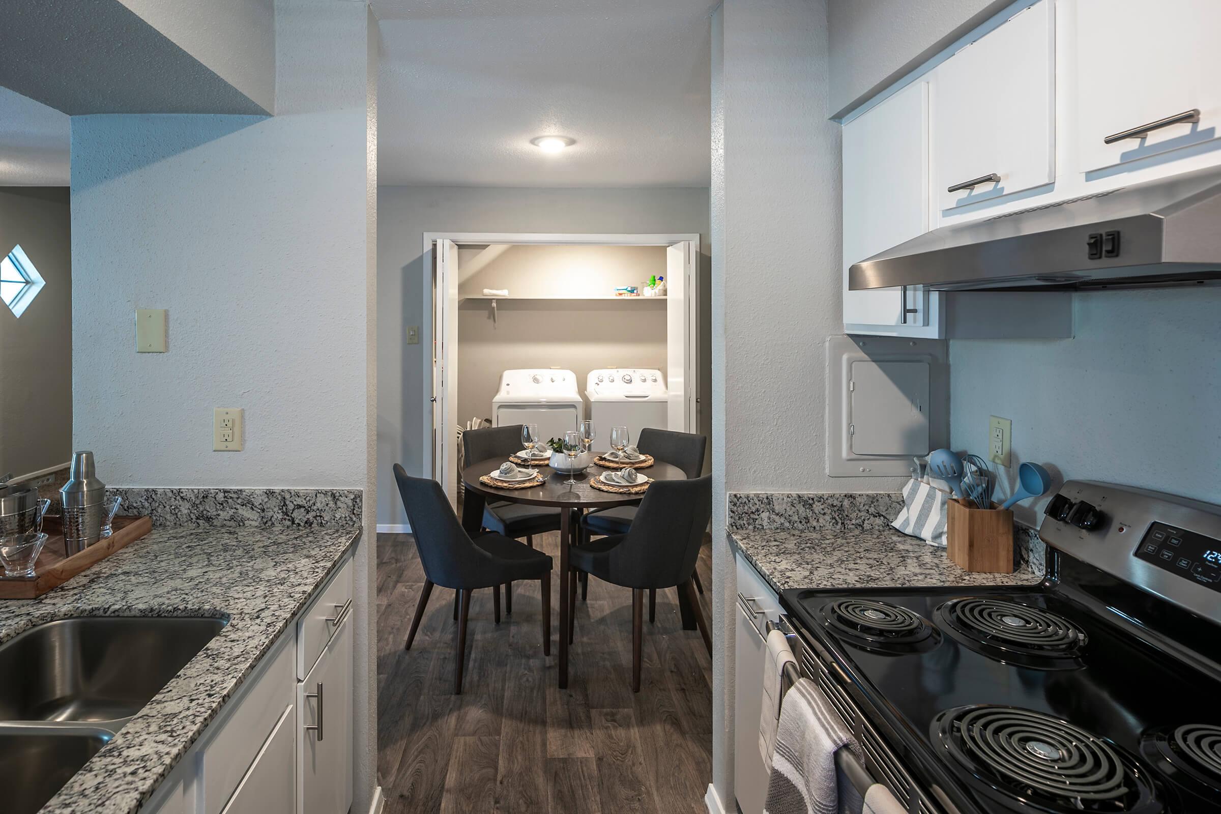 Modern kitchen featuring granite countertops, stainless steel appliances, and a stovetop. A small dining area with a round table set for two is visible, along with a laundry area in the background. The space has neutral-colored walls and stylish chairs, creating a cozy atmosphere.