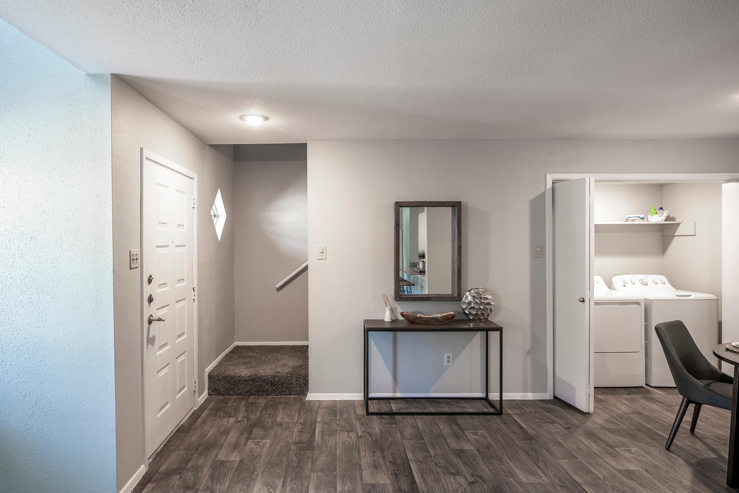 A modern interior view of a small entryway featuring a front door, a staircase leading up, and a laundry room behind sliding doors. The space includes a decorative mirror above a console table and a simple chair. The flooring is a dark wood laminate, and the walls are painted in soft, neutral colors.