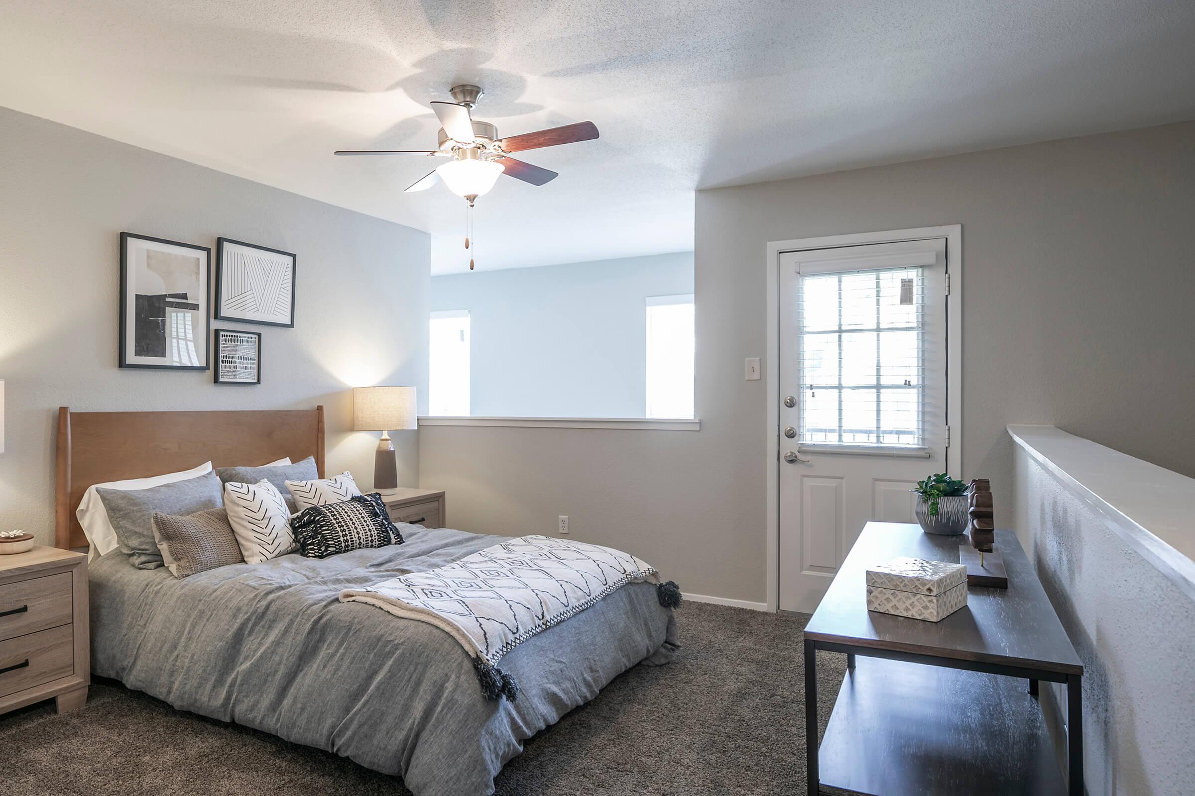 A cozy bedroom featuring a large bed with gray bedding and decorative pillows. There's a wooden nightstand with a lamp beside the bed, and a dark wooden console table against the wall. A ceiling fan hangs overhead, and a door with a window leads outside. Soft gray walls create a calm atmosphere.