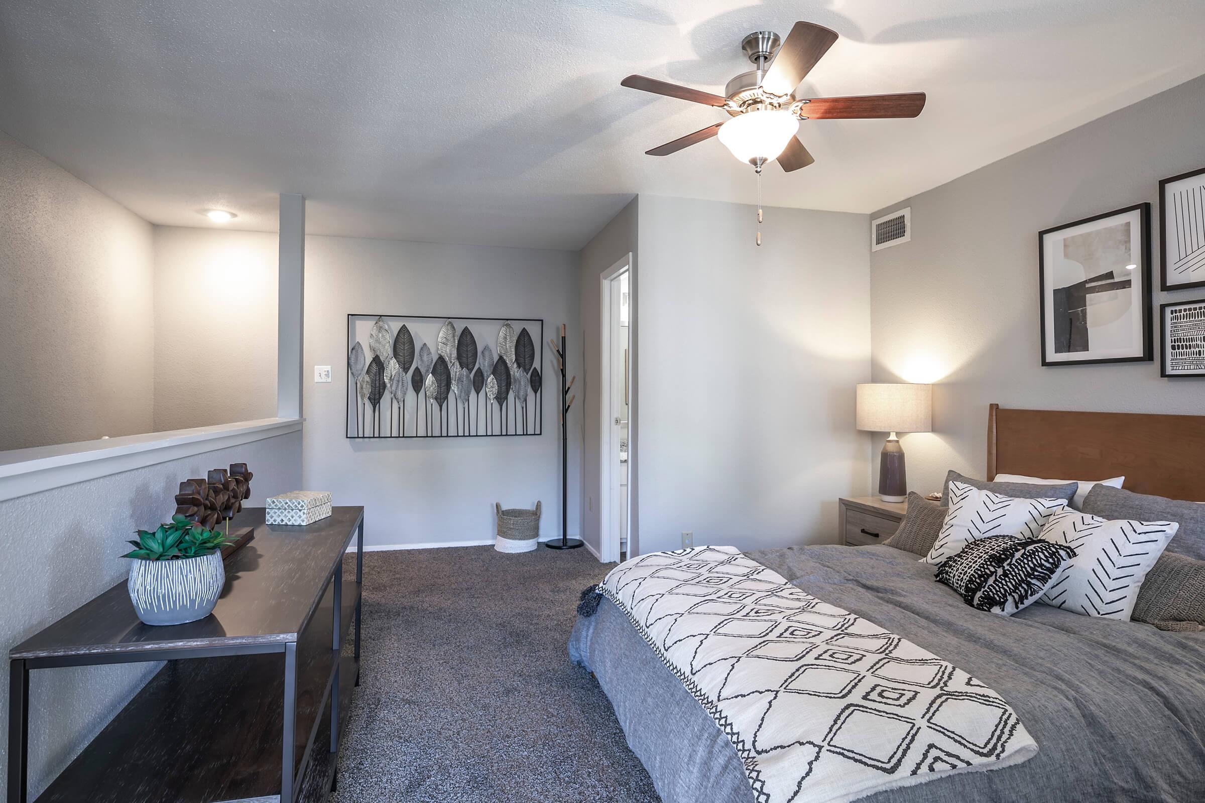 A cozy bedroom featuring a textured gray wall, a king-sized bed with decorative pillows, a stylish lamp on a nightstand, and a ceiling fan. A small decorative table with a plant adds to the ambiance, while framed artwork and a subtle decor create a serene atmosphere.