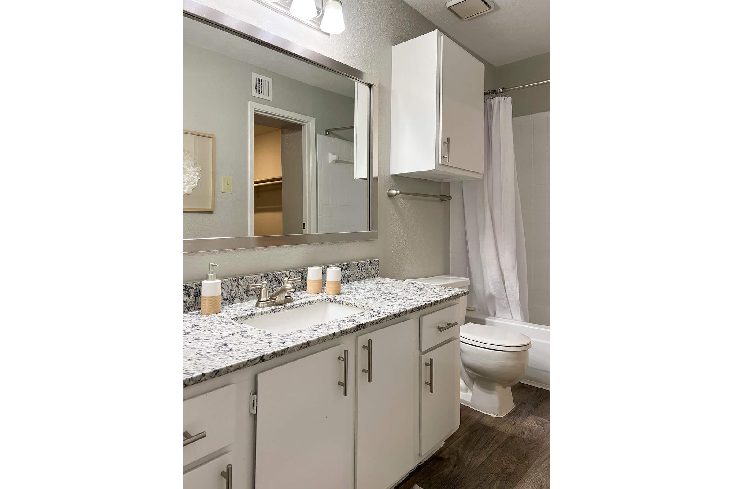 A modern bathroom featuring a white granite countertop with dual sinks, a large mirror above, and white cabinetry. A shower with a white curtain is visible. The flooring is a dark wood, and there's a small cabinet on the wall. The overall decor is clean and contemporary.
