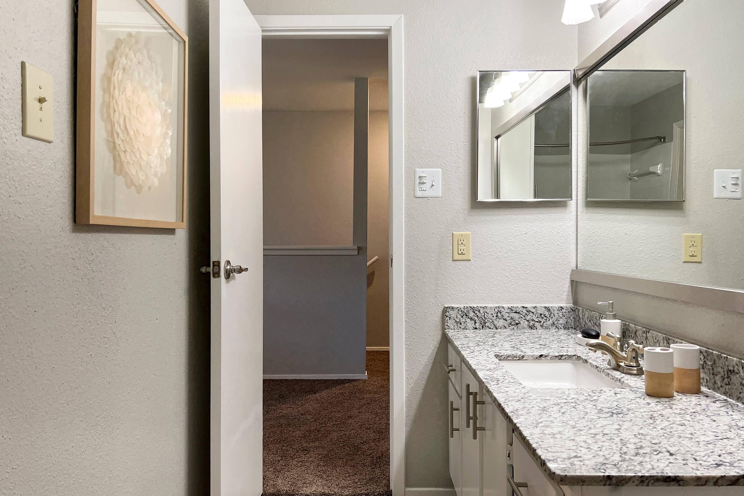 A well-lit bathroom featuring a granite countertop with a sink and modern fixtures. A mirror above the sink reflects the space, and a doorway leads to a hallway with carpeted flooring. A framed artwork is on the wall near the door, adding a decorative touch to the otherwise simple and clean design.
