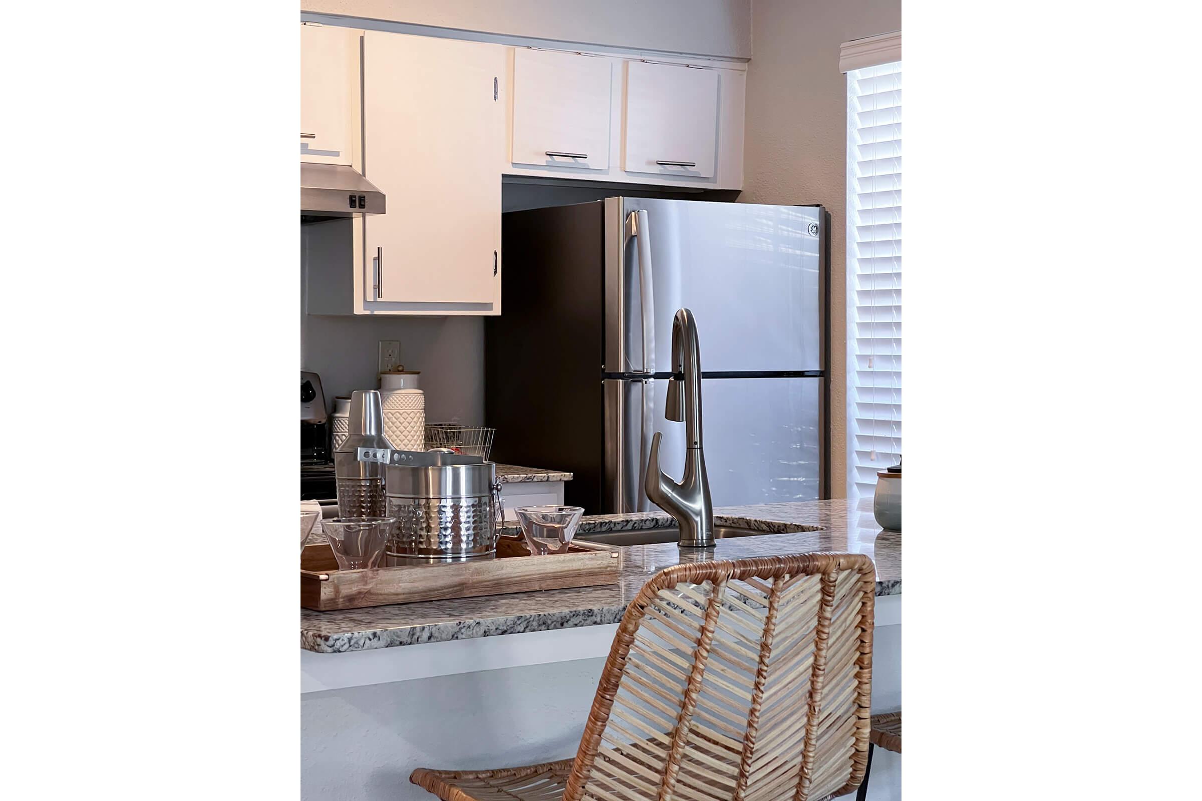 Modern kitchen featuring white cabinets, a sleek stainless steel refrigerator, and a countertop with a wooden tray holding glassware. A woven dining chair is positioned at the counter, with natural light streaming through a window adorned with horizontal blinds.