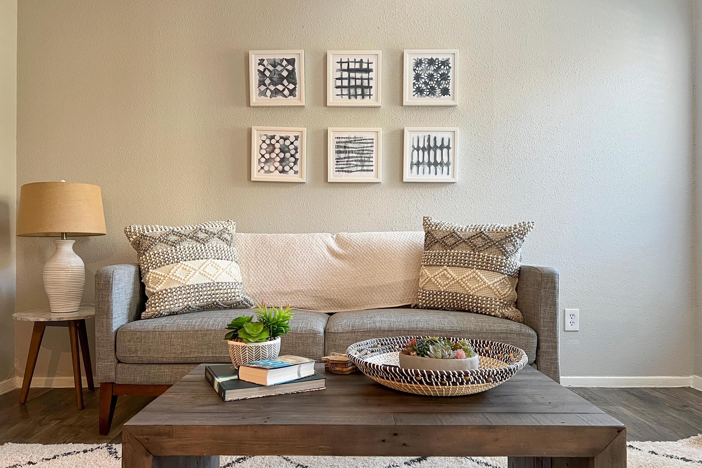 A cozy living room featuring a gray sofa with decorative pillows, a wooden coffee table with a plant and books, and a textured basket. On the wall above the sofa, six framed black-and-white artworks create a modern aesthetic. A lamp on a side table adds warmth to the space.