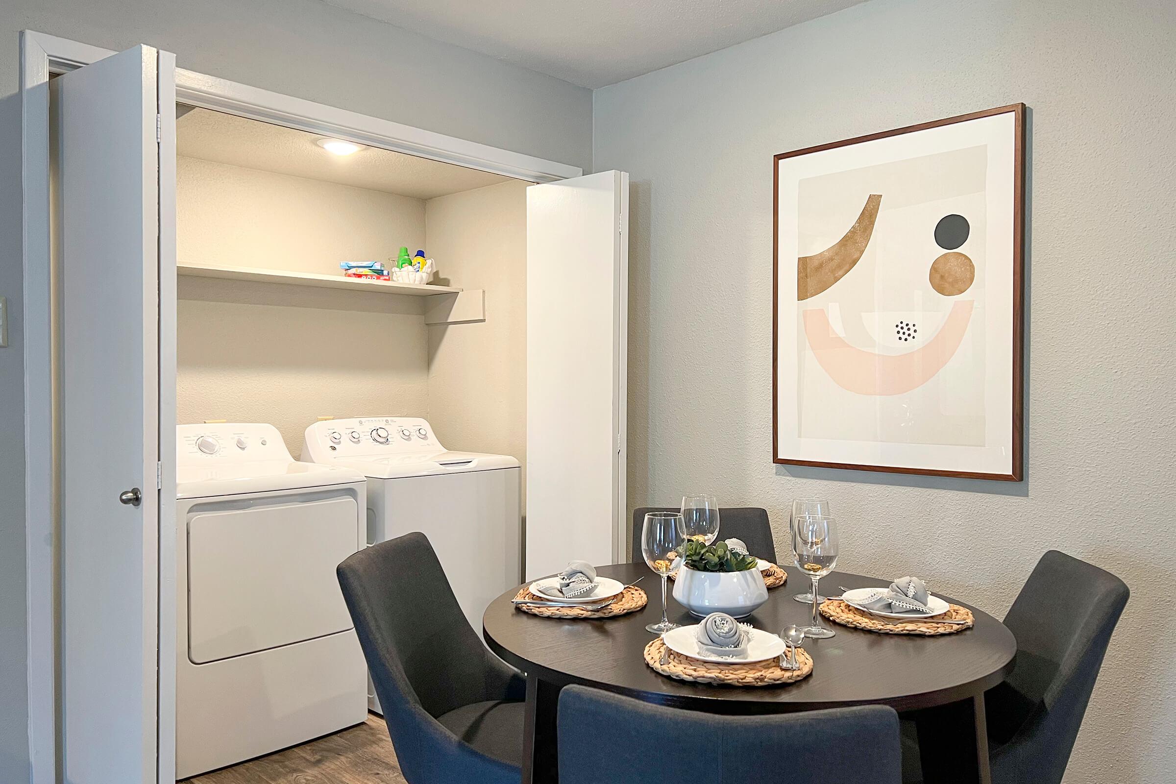 A bright, modern dining area featuring a round table set with dishes, wine glasses, and a centerpiece. In the background, there is a laundry area with white washing machines visible through an open door, and a framed abstract artwork hangs on the wall. The room has neutral colors, enhancing its spacious feel.
