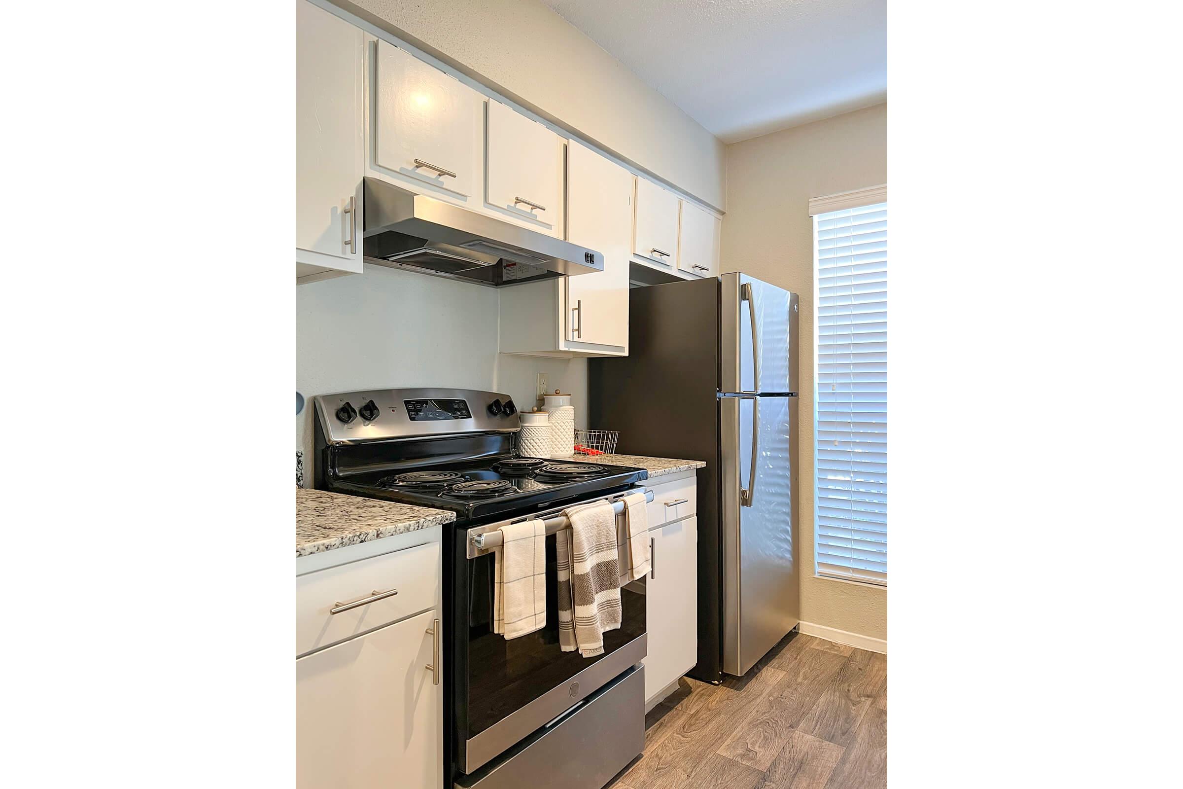 A modern kitchen featuring a stainless steel refrigerator and oven, white cabinets, and a granite countertop. The stove has a pot and kitchen towels hanging from it, with natural light coming through a window. The flooring is a light wood, enhancing the contemporary design.