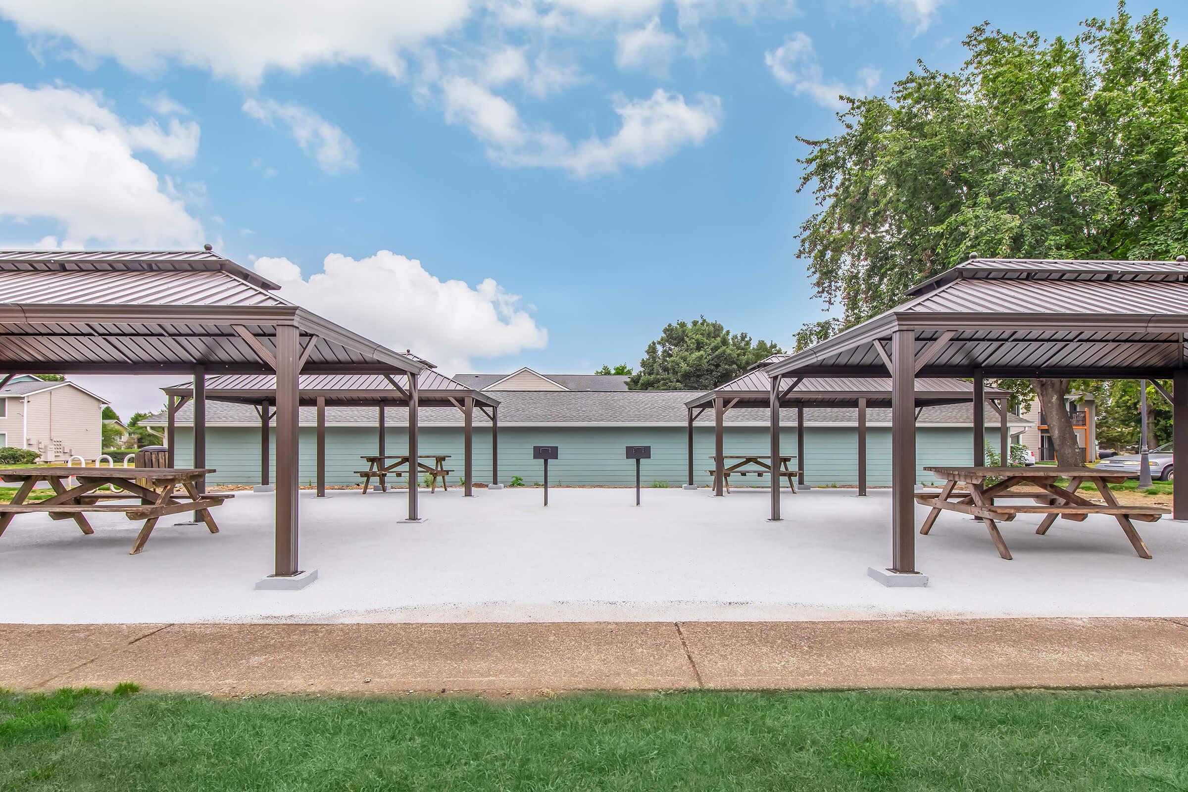A spacious outdoor pavilion area featuring multiple covered picnic tables and grills, with a smooth concrete floor and green grass surrounding the area. The background includes trees and residential buildings under a partly cloudy sky.