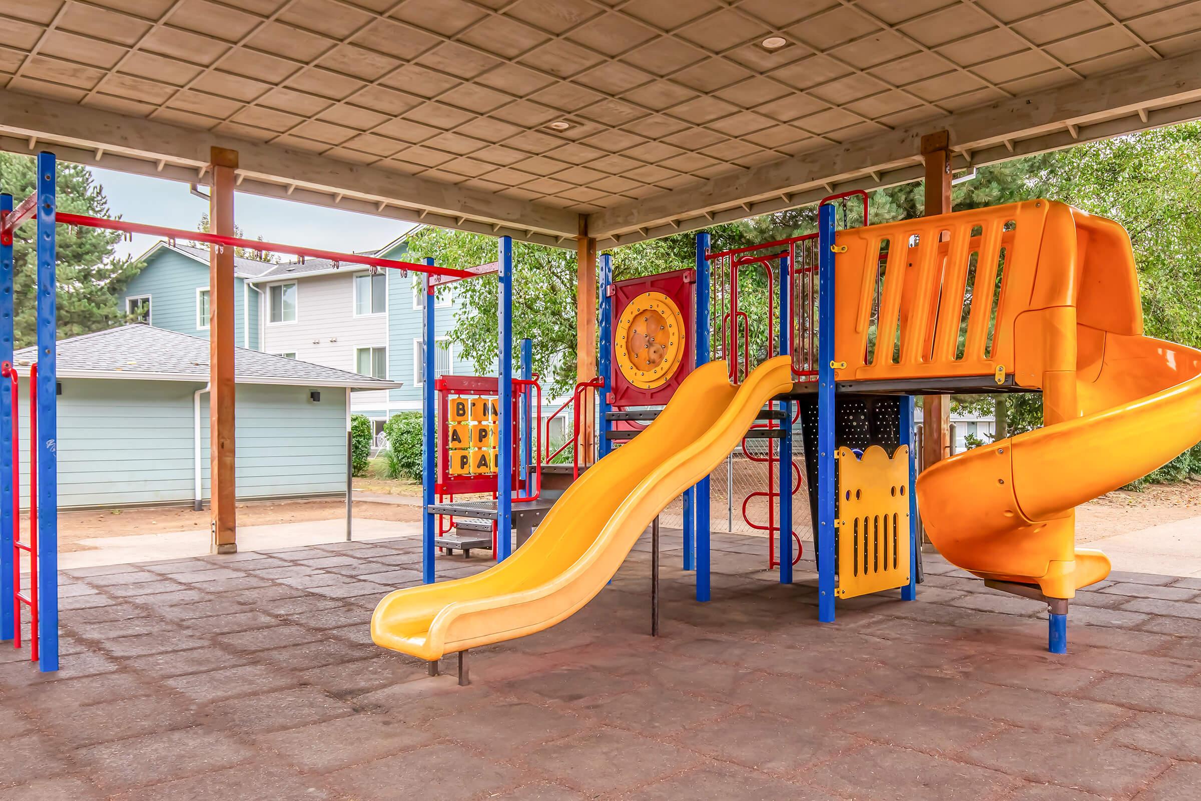 A colorful playground structure featuring a tall orange slide, climbing areas, and various interactive panels, set under a covered area. Nearby residential buildings are visible in the background, with green trees and an open space around the playground.
