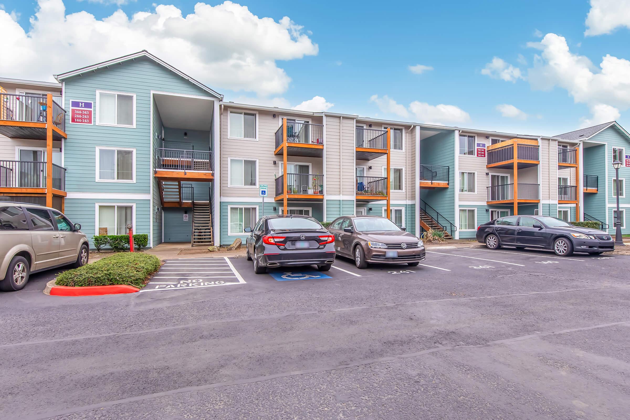 A multi-story apartment building with light blue and gray siding. The building features balconies with orange railings. Several parked cars are visible in the foreground, along with a landscaped area. The sky is partly cloudy, creating a bright, inviting atmosphere.
