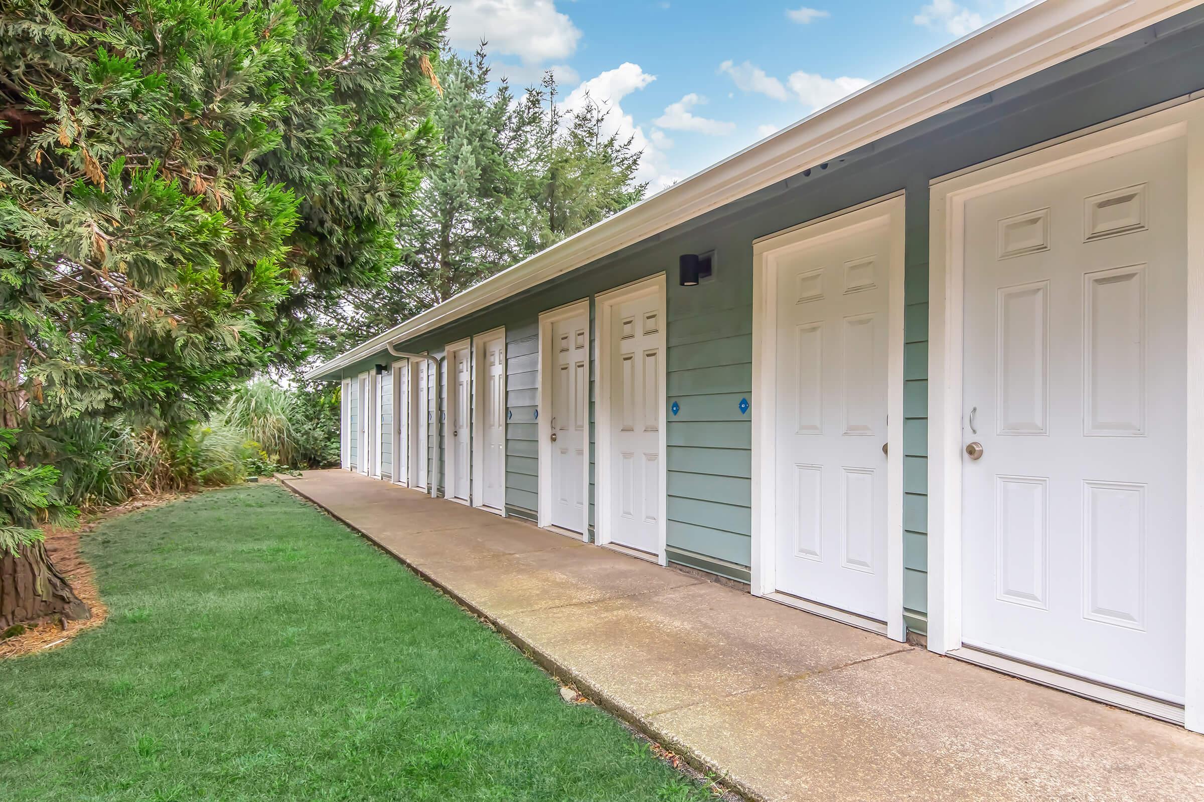 A pathway leading to several identical white doors on a light blue building, surrounded by green grass and trees. The sky is partly cloudy, creating a serene outdoor atmosphere.