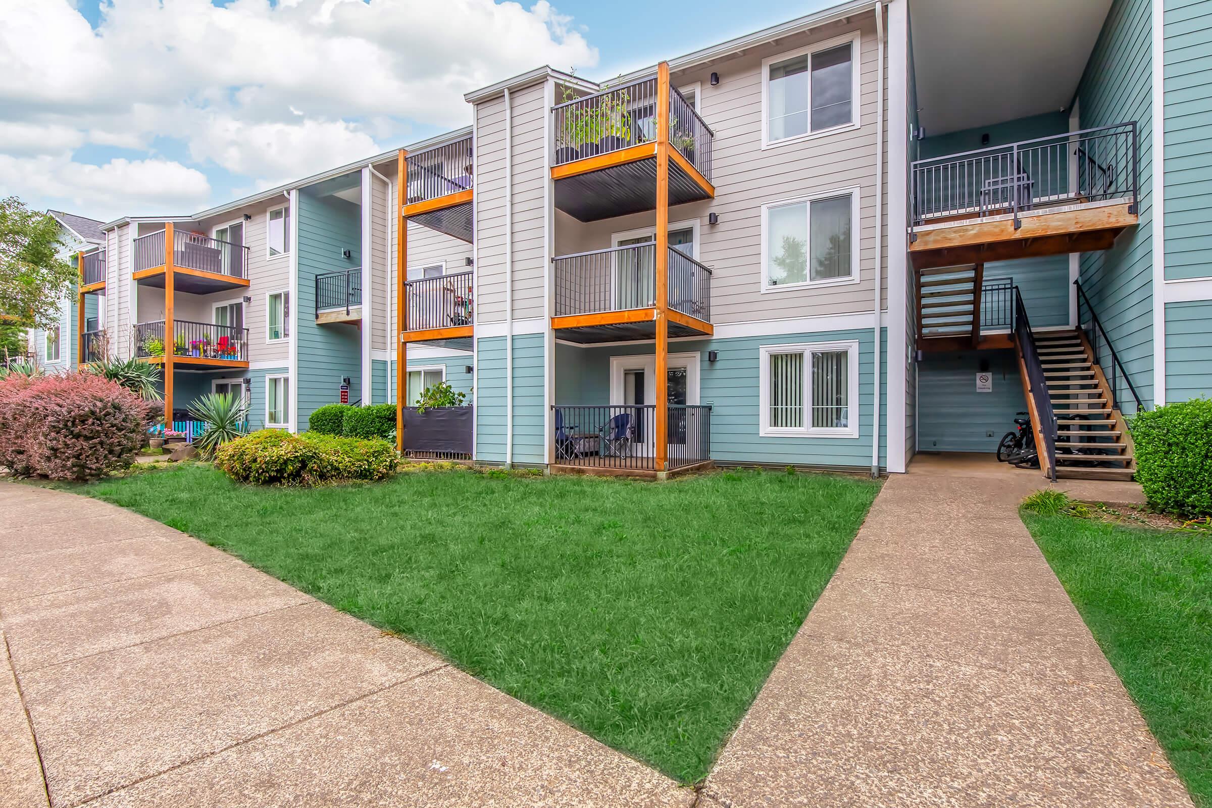 A view of an apartment complex featuring multiple balconies with wooden railings. The building has light blue siding and a well-maintained lawn with a pathway. Surrounding plants and shrubs enhance the greenery, and one staircase leads to the upper level of the complex.