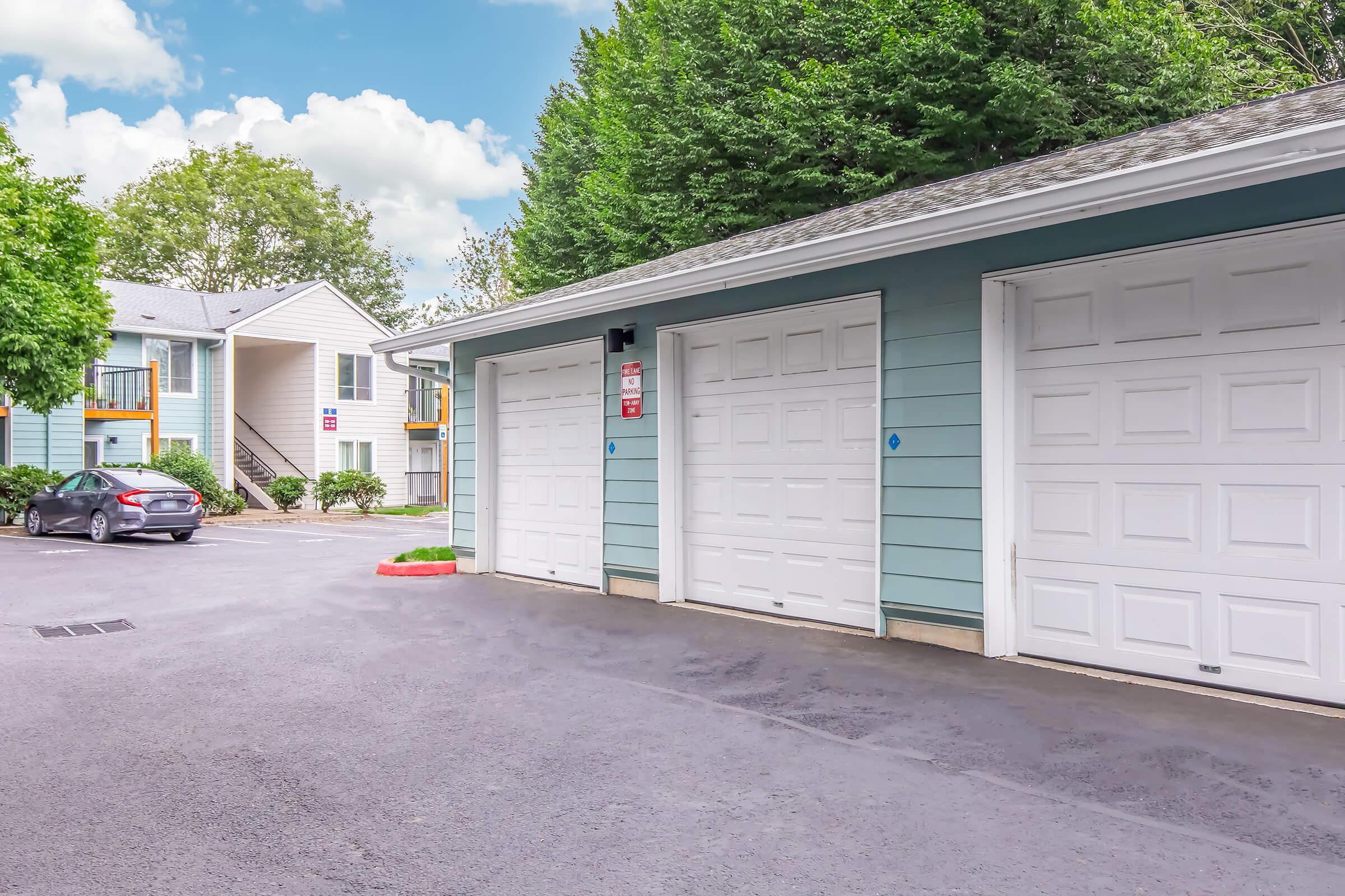 A parking area featuring several garages with white doors, surrounded by green trees and a light blue sky. In the background, there is a building with balconies and a car parked nearby. The scene conveys a residential atmosphere.