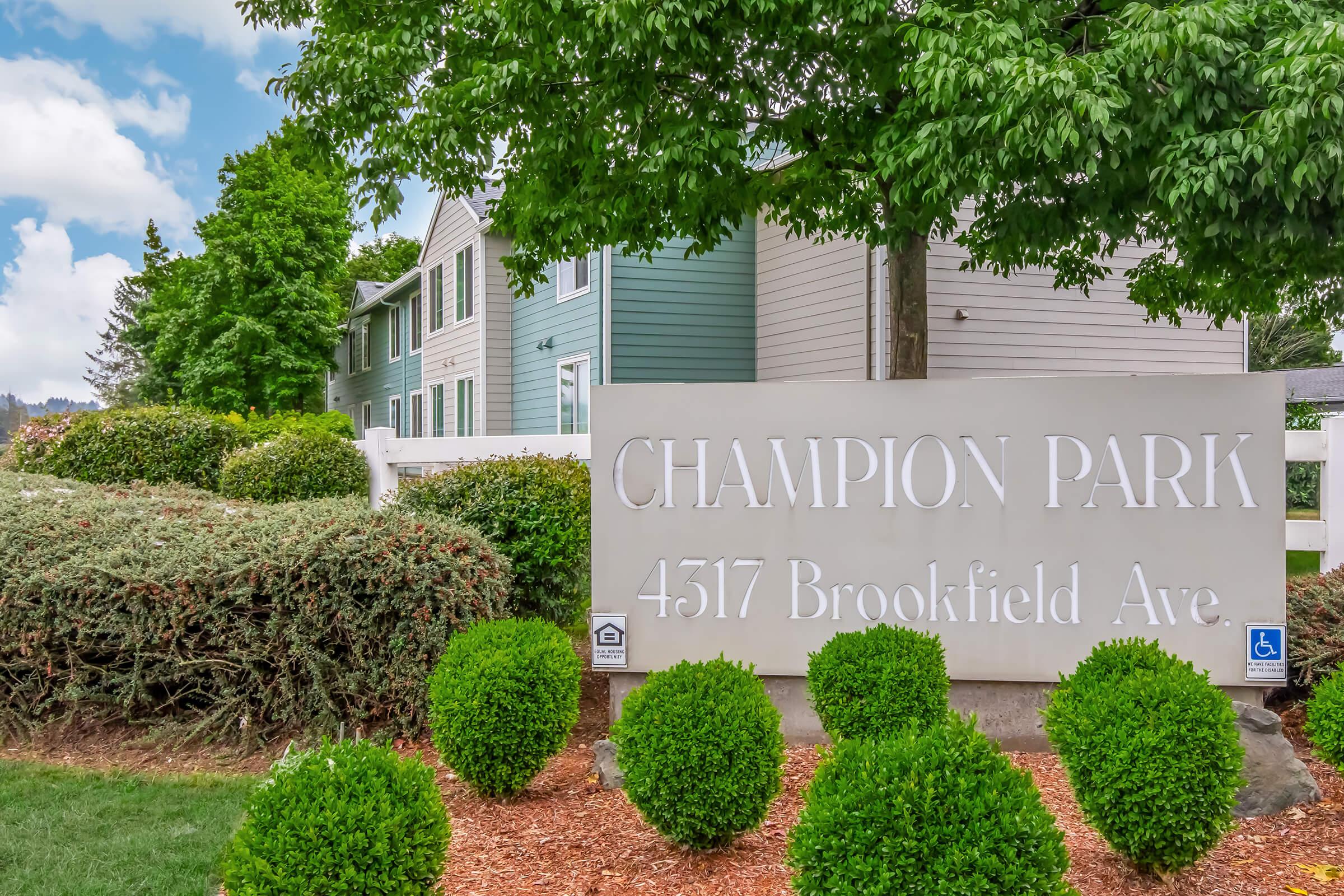 Sign for Champion Park with the address "4517 Brookfield Ave" in a landscaped area featuring neatly trimmed bushes and trees, set against a backdrop of residential buildings. The sign is prominent and modern, showcasing the name of the property.