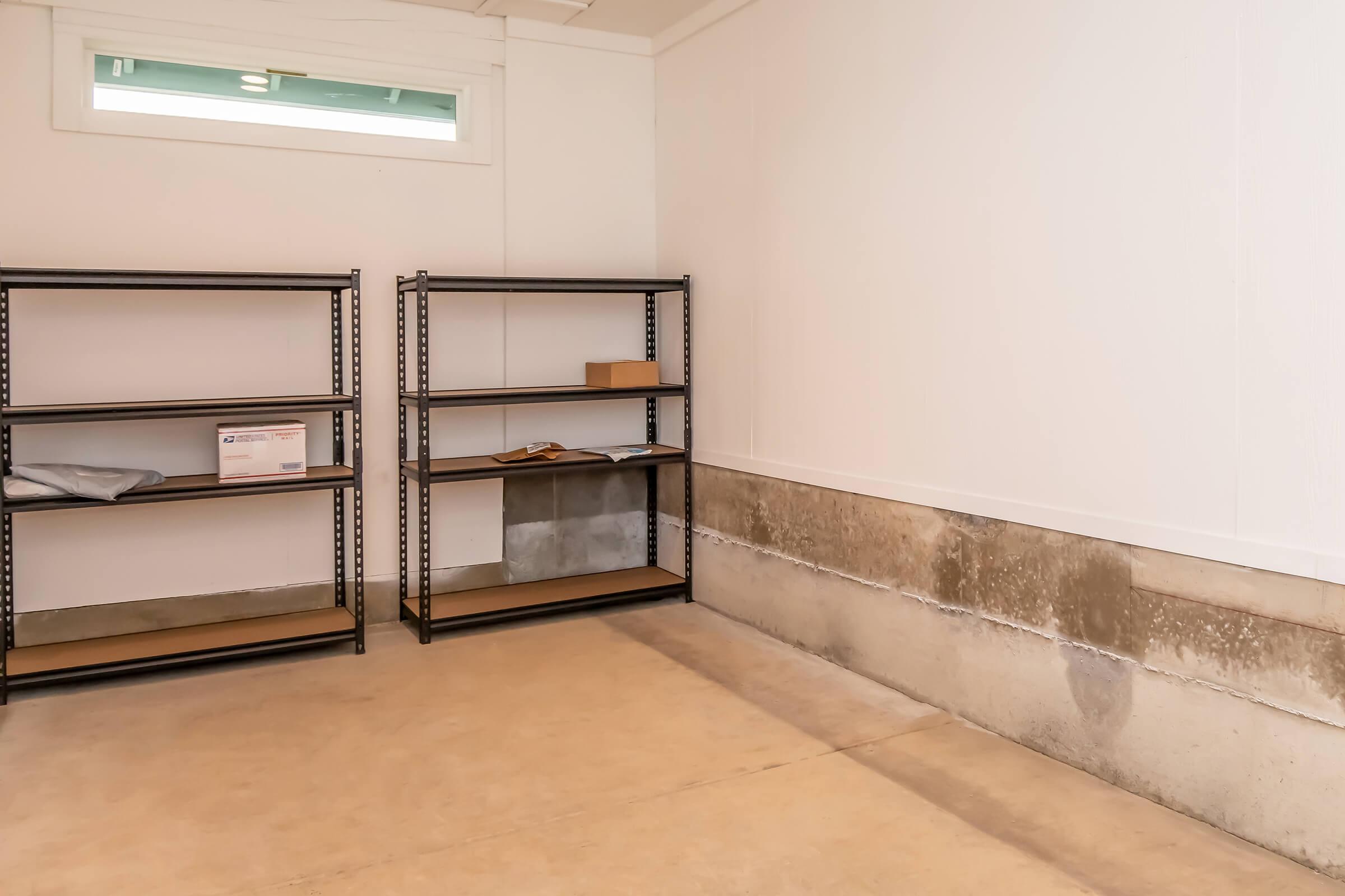 An empty room with white walls and a concrete floor. On the left, there are two metal shelving units with wooden shelves, one partially filled with boxes. The far wall features a plain, unadorned surface with a low concrete ledge. A small window is visible near the ceiling, allowing light to enter the space.