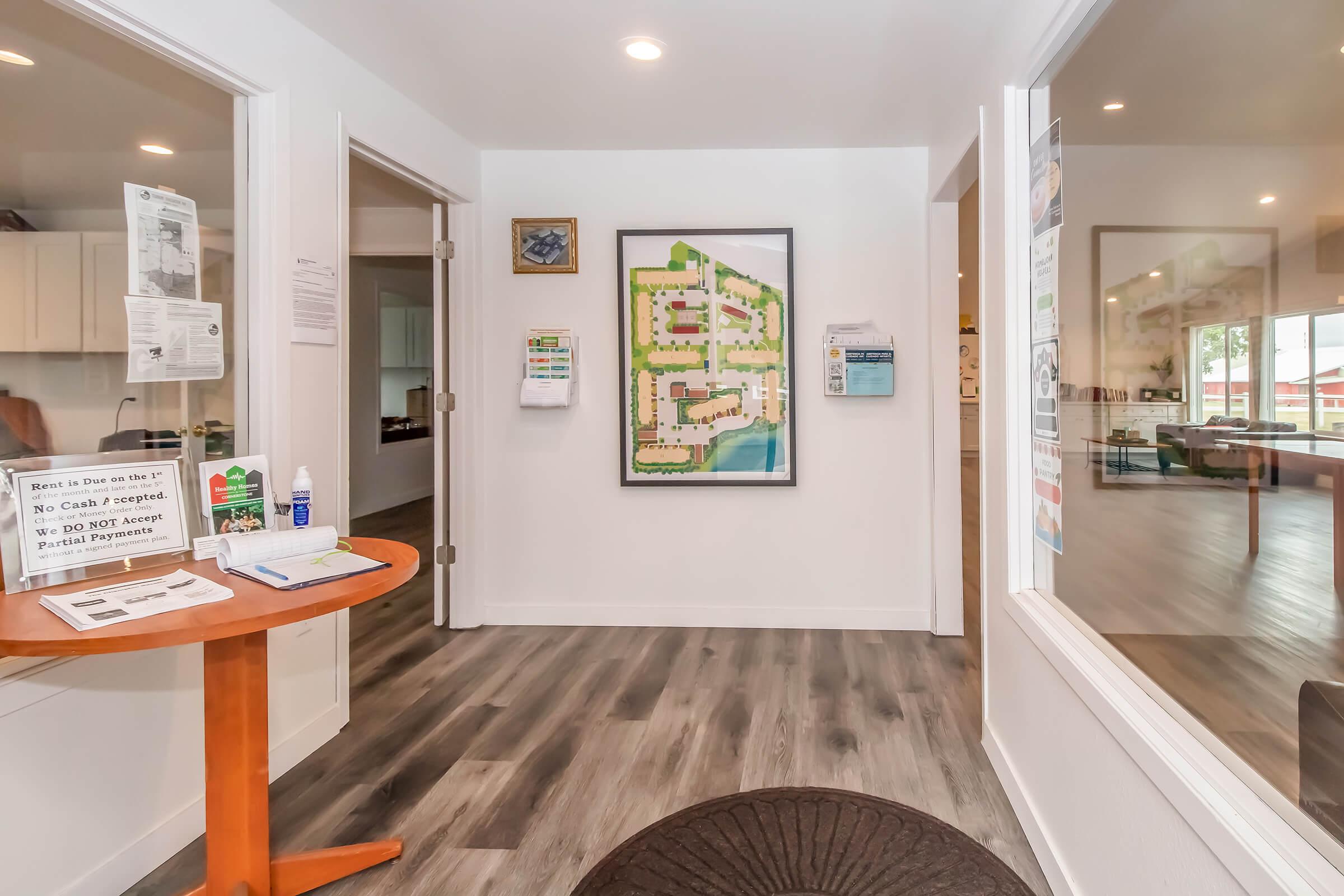 Interior view of a welcoming reception area featuring a round wooden table with informational materials, a colorful map on the wall, and clear pathways leading to other rooms. Natural light enters through large windows, creating a bright and open atmosphere.