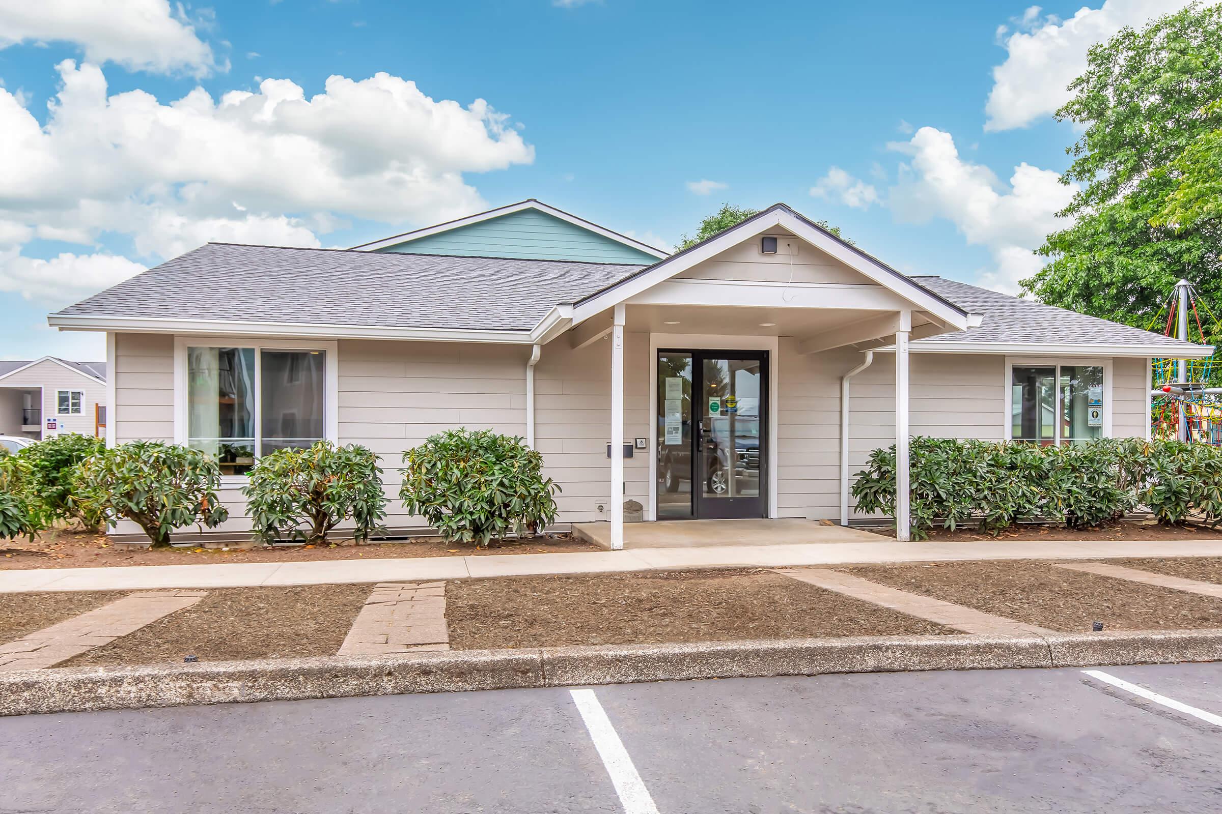 A single-story office building with a light-colored exterior and a sloped roof. It features large windows, a central entrance with glass doors, and landscaped greenery along the front. In the background, there is a playground visible, and the sky is partly cloudy, suggesting a bright day.