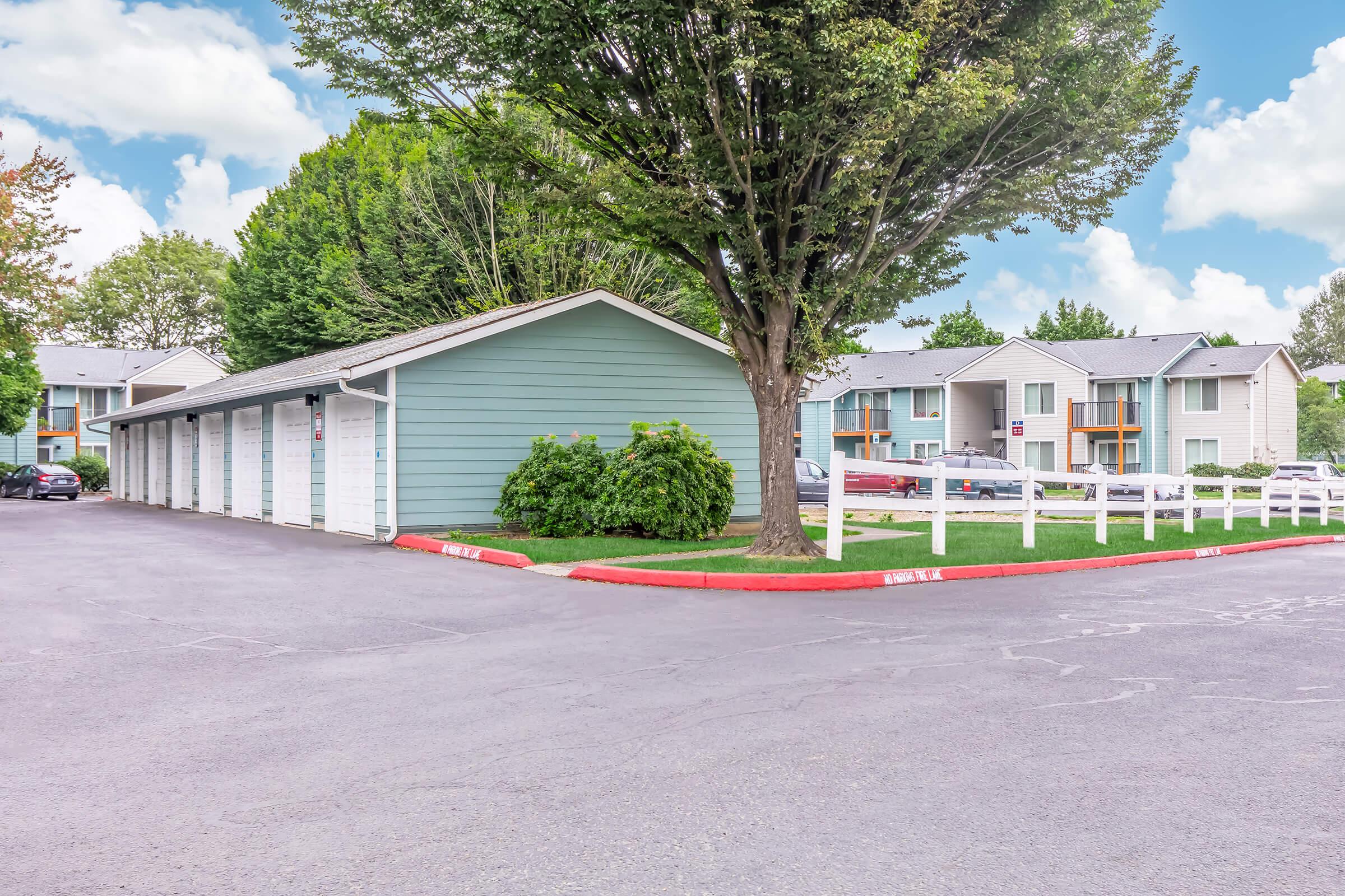 A view of a residential complex featuring a light blue storage building next to a well-manicured lawn and trees. In the background, there are multi-unit apartments with balconies. The parking area has a smooth asphalt surface and is lined with a white fence. Bright sky with scattered clouds.