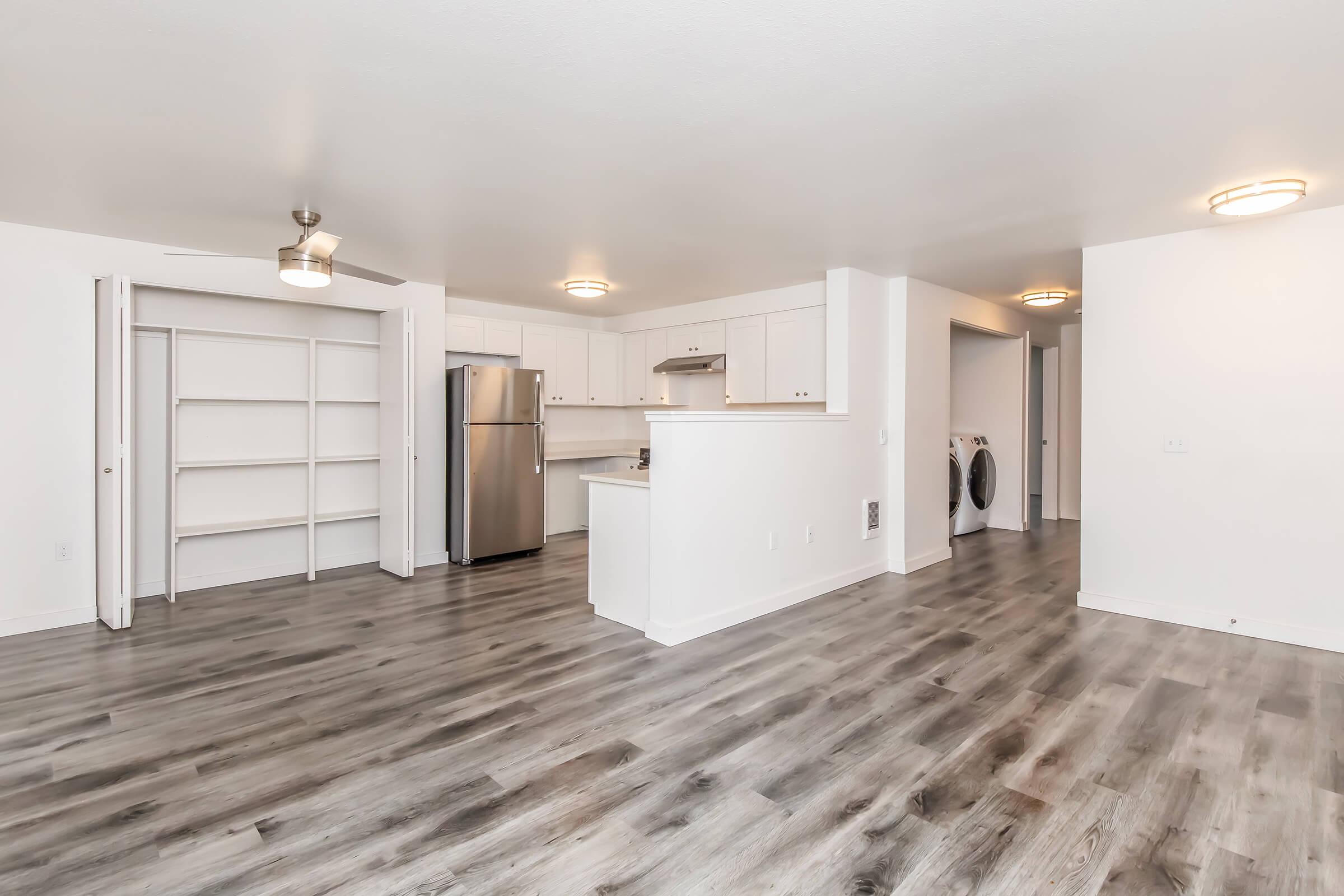 A modern, open-concept living space featuring a kitchen with stainless steel appliances, a large island, and light-colored cabinetry. The flooring is a light wood laminate. There are built-in shelves on one wall and laundry appliances visible in the background, along with ample natural light from ceiling lights.
