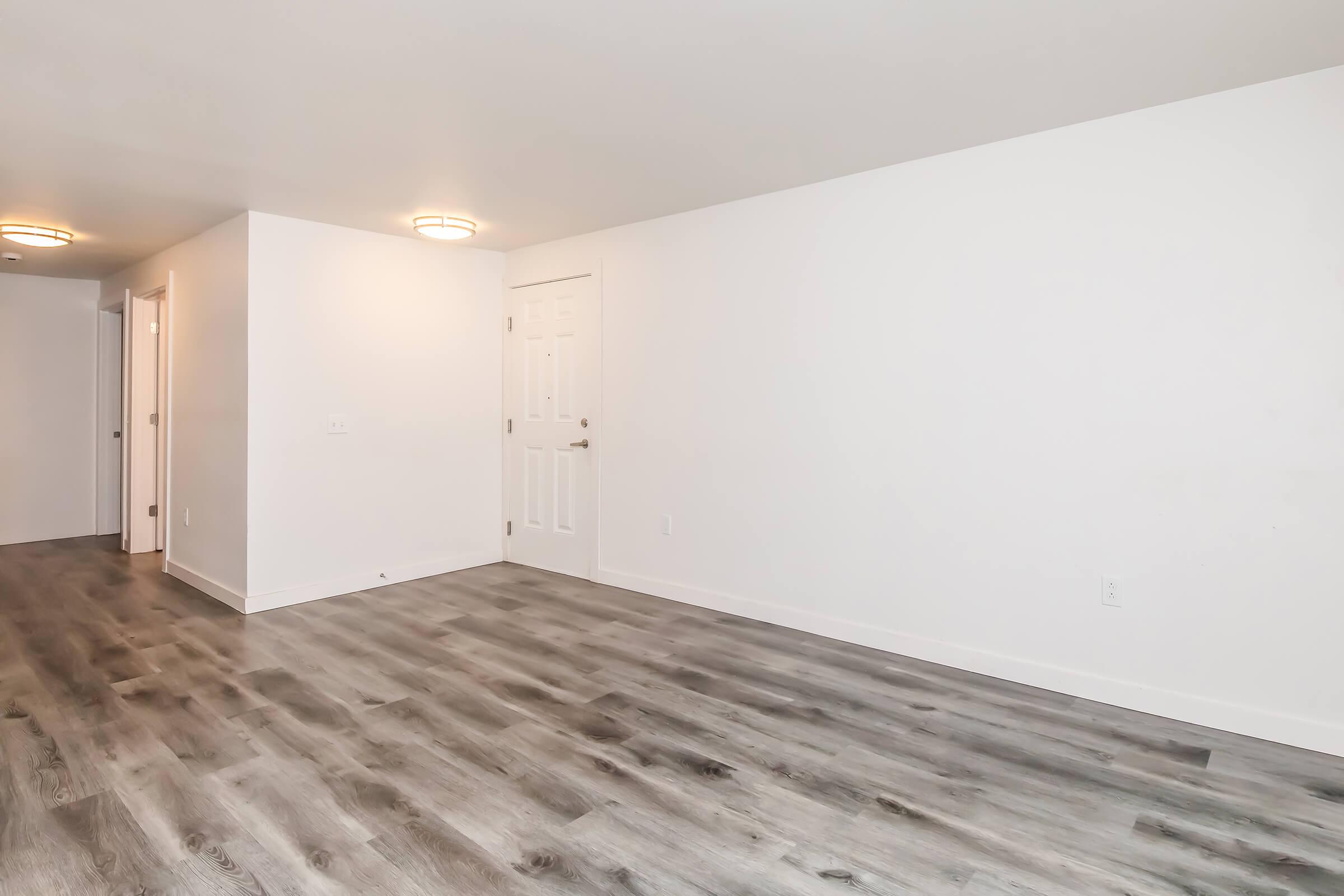 A clean, empty room with light gray laminate flooring. The walls are white, and there are two ceiling lights providing illumination. A closed door is visible on the right, while an open doorway leads to another room on the left. The space has a minimalist feel, suitable for various furnishings or decor.