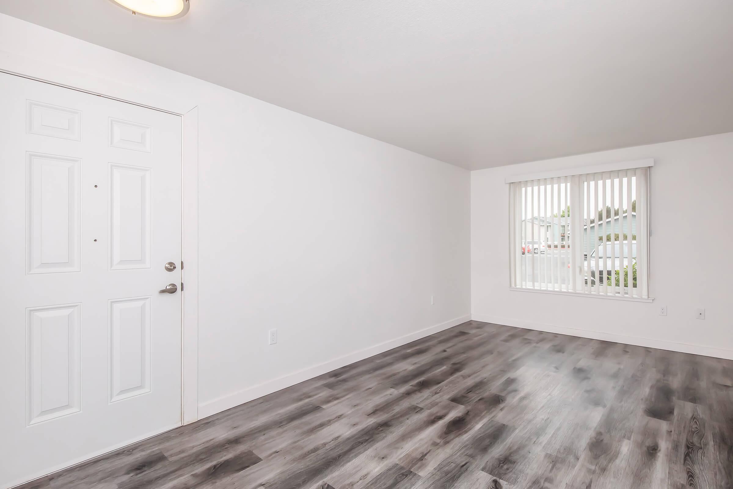 Bright and empty room with light gray walls, modern laminate flooring, and vertical blinds on a window. The room includes a front door with a simple design, emphasizing a clean and minimalist aesthetic.