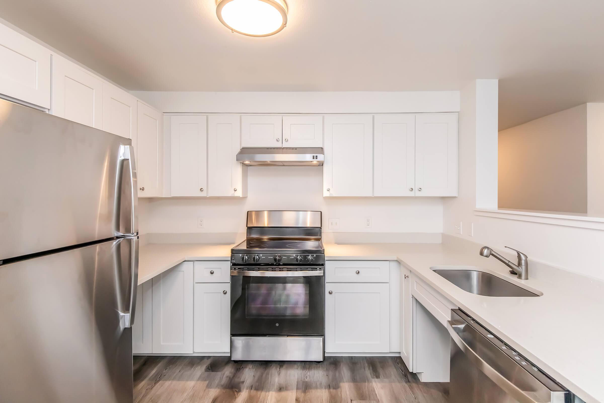 A modern kitchen featuring white cabinetry, a stainless steel fridge, a gas stove with an overhead exhaust, and a stainless steel sink. The countertops are light and the flooring is wood-like, creating a clean and contemporary look. Bright lighting enhances the overall appeal of the space.