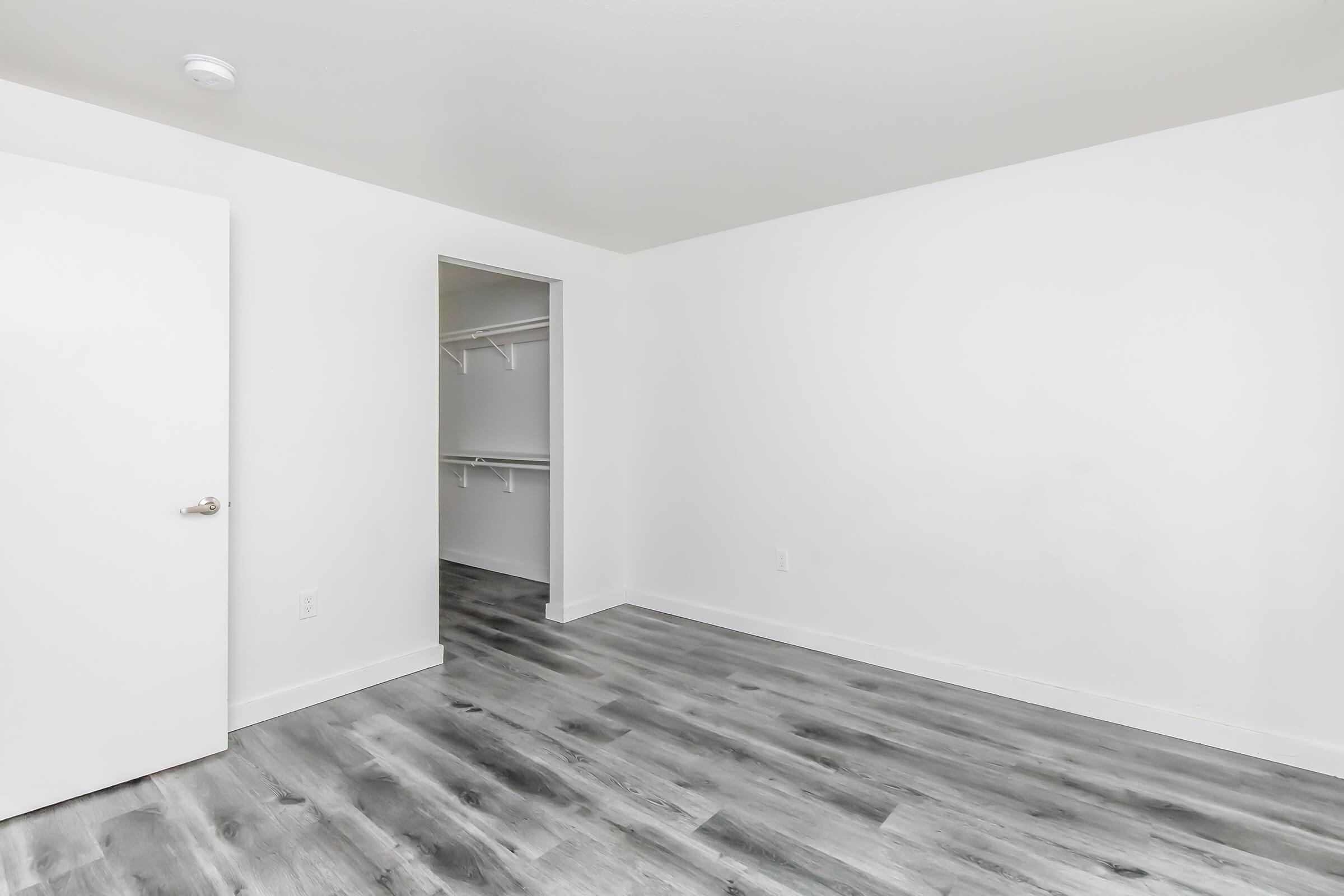 A spacious, empty room featuring light gray vinyl flooring and white walls. There is a closed white door on the left and a closet entrance on the right with hanging space. The room is bright and uncluttered, creating a minimalist aesthetic.