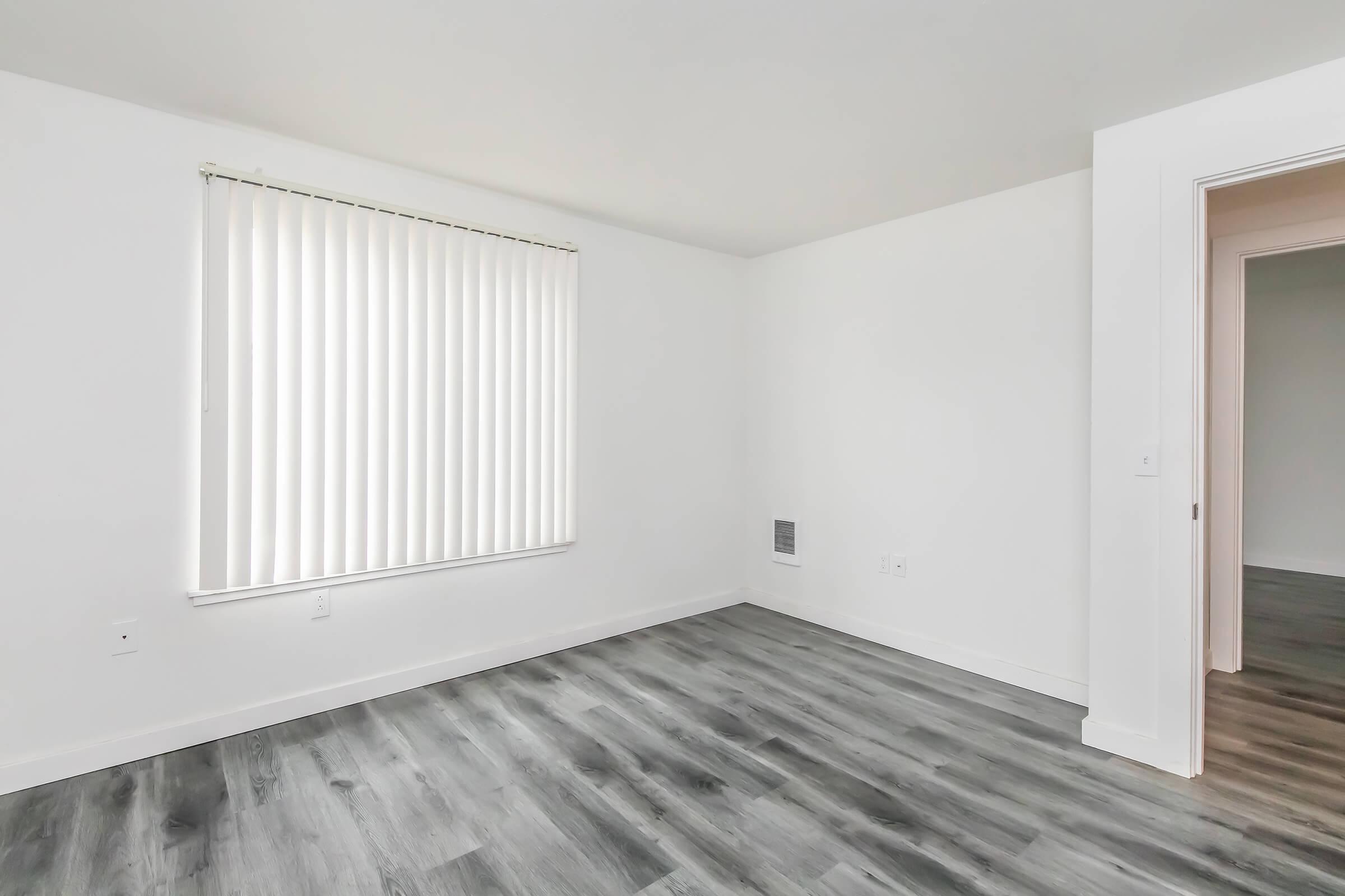 Empty room with light-colored walls and wood-like flooring. A large window with vertical blinds provides natural light, and a door on the right leads to another space. The room has a clean, minimalist design, creating an open and airy atmosphere.