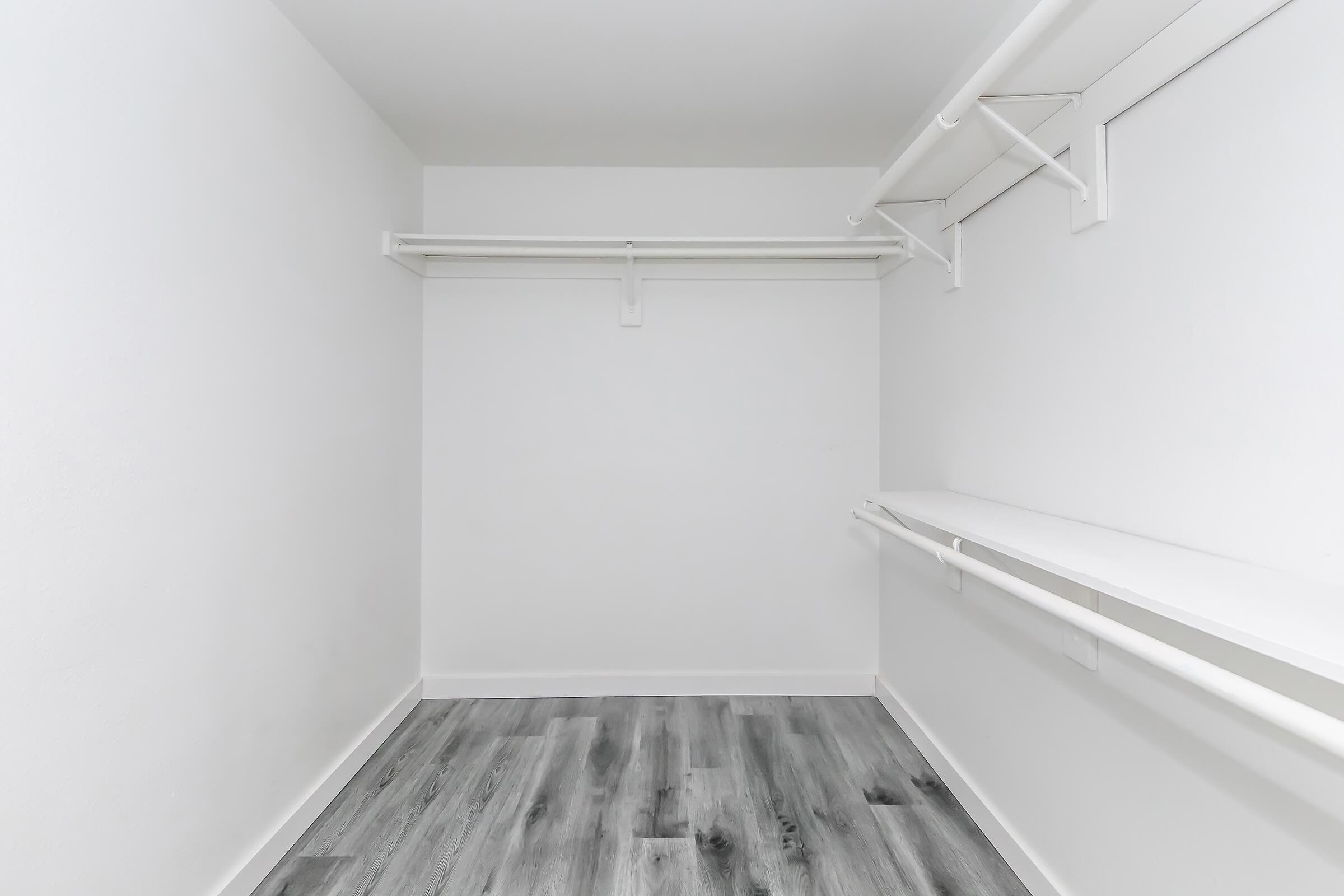 Empty closet with white walls, a light gray wooden floor, and two hanging racks along the back wall. There is a shelf along one side, creating a clean, minimalistic space suitable for storage or organization.