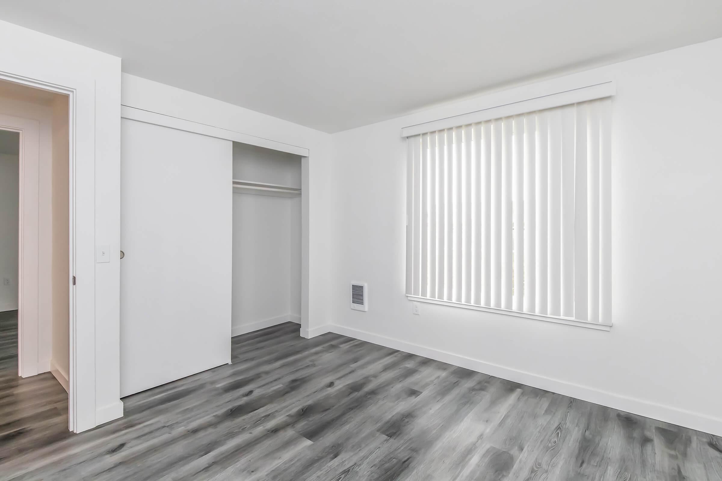 A bright, empty room with white walls and a large window covered by vertical blinds. There is a closet with sliding doors on the left. The floor features gray laminate, creating a modern and spacious feel. The overall design is minimalistic and clean, suitable for various decorating styles.