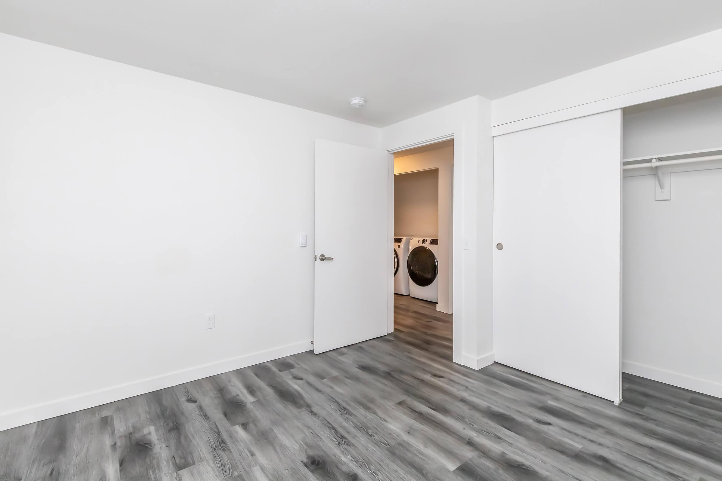 Empty room with light-colored walls and gray wooden flooring. Two white doors are visible: one closed and one slightly ajar, leading to a laundry area with washing machines. The room is well-lit and uncluttered, creating a spacious feel.