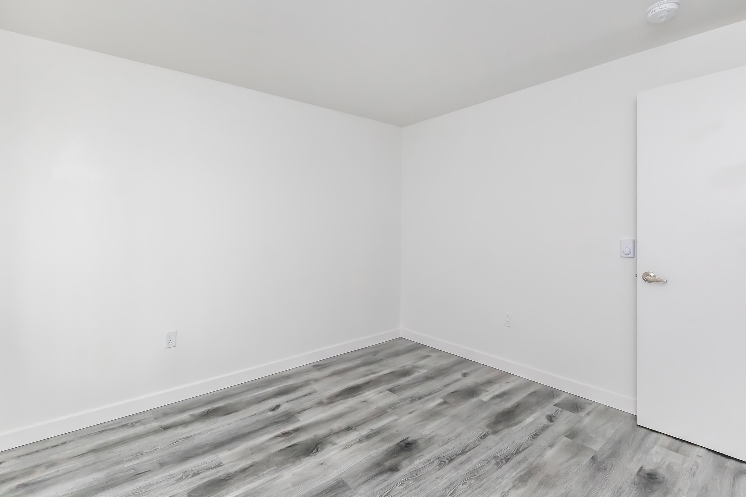 Empty room with white walls and a light gray laminate floor. A closed white door is visible on the right side, and the space appears bright and uncluttered, suggesting a minimalist design. No furniture or decorations are present.