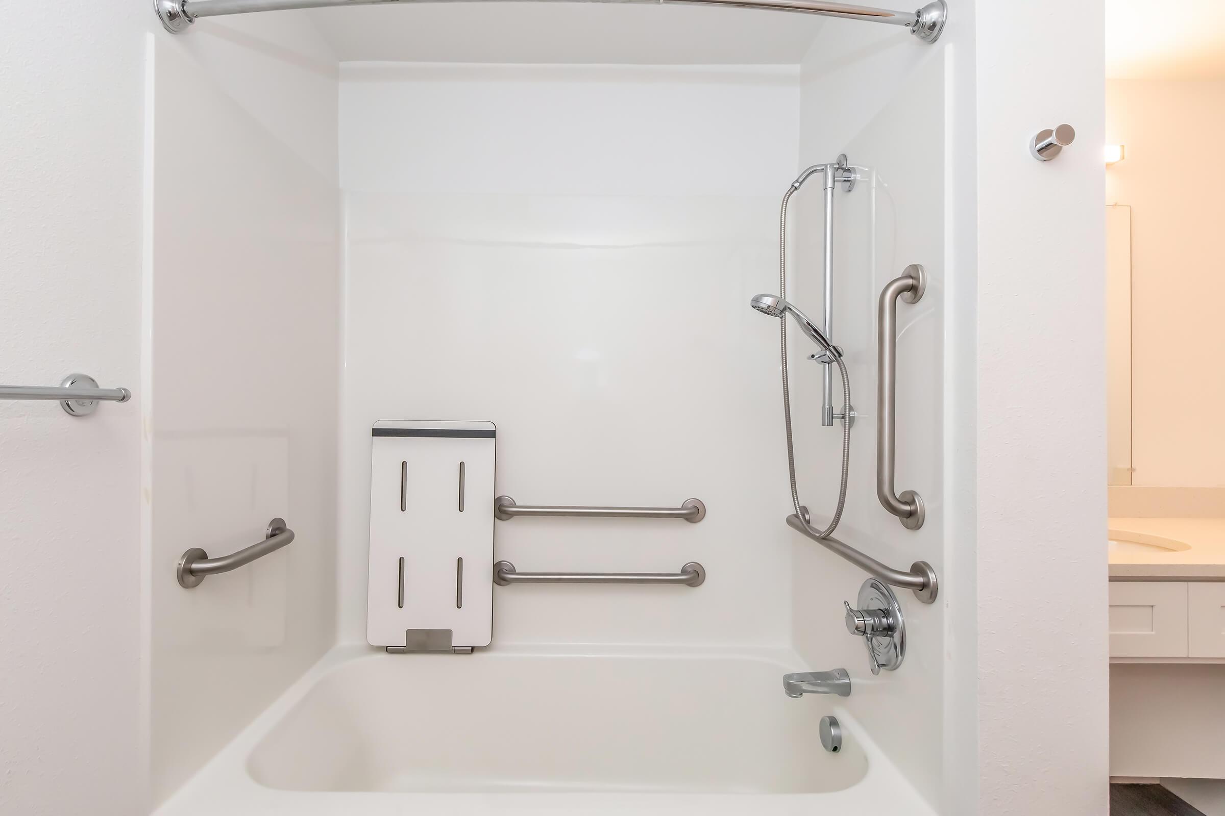 Accessible shower stall with a built-in seat, grab bars on the walls, and a handheld showerhead. The walls are plain white, and the tub area is designed for safety and assistance for individuals with mobility challenges. A modern bathroom counter is visible in the background.