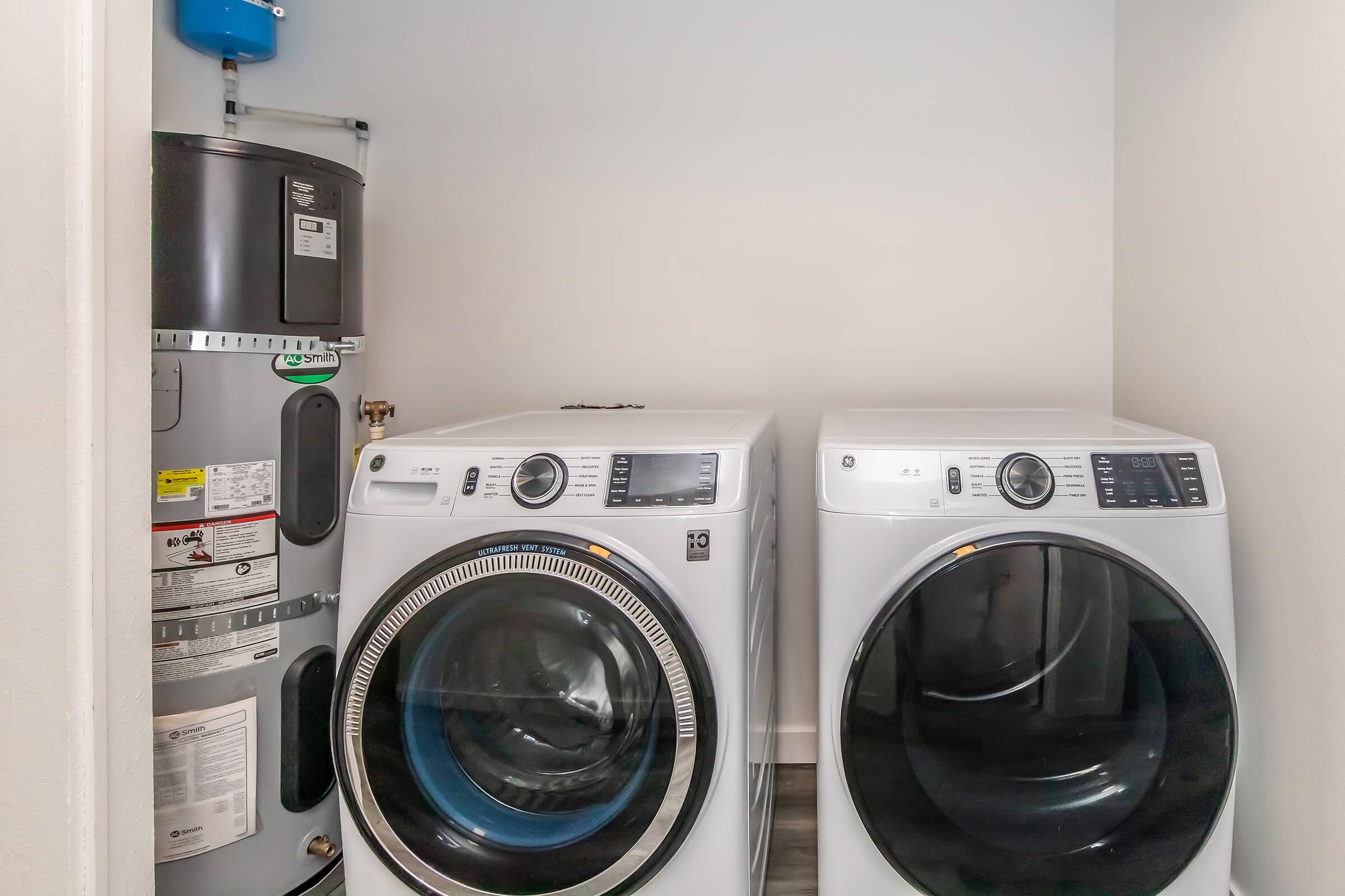A small laundry room featuring two white front-loading washing machines side by side, with a gray water heater positioned next to them. The walls are painted white, and the floor has a light-colored tile. The space appears tidy and well-organized.