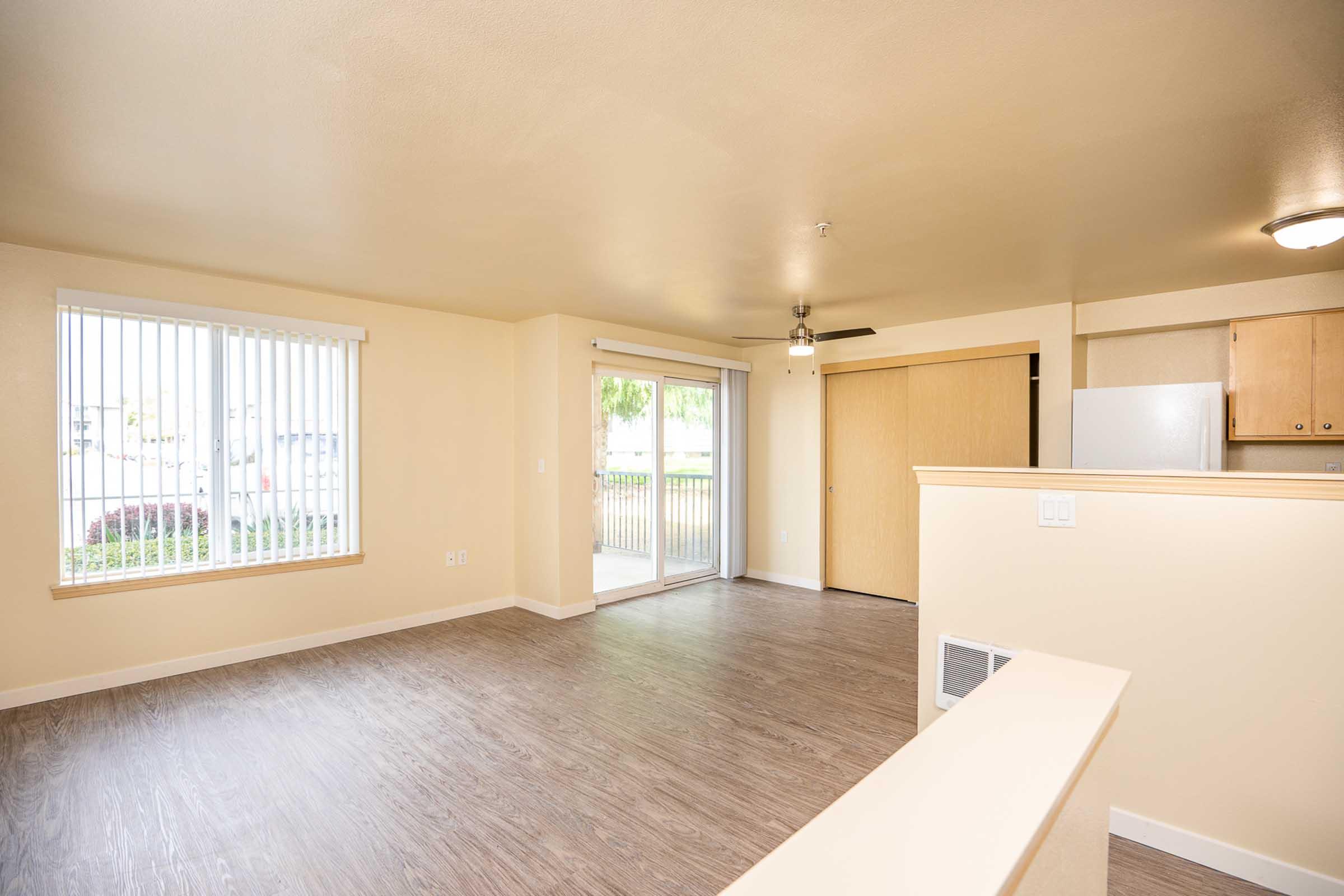 A spacious, light-colored living area with a sliding glass door leading to a patio. There are large windows with vertical blinds, a ceiling fan, and laminate flooring. The kitchen area is partially visible, featuring wooden cabinets and a refrigerator. The walls are painted a soft beige.