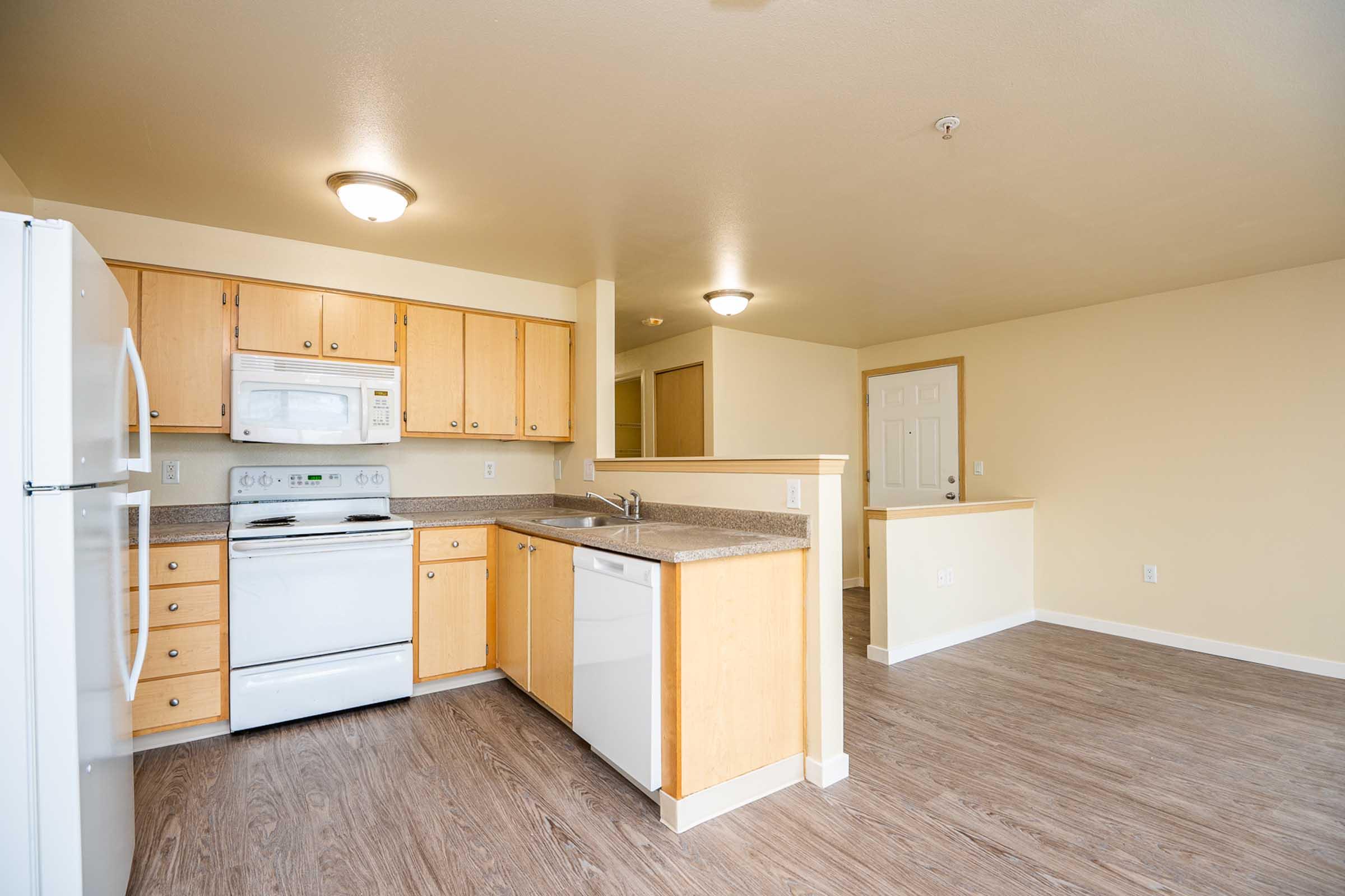 Modern kitchen with light wood cabinetry, white appliances including a refrigerator, microwave, and stove. The countertop is gray and there is a dishwasher. Open layout leads to a brightly lit living area with neutral walls and hardwood-style flooring. Soft overhead lighting enhances the space.