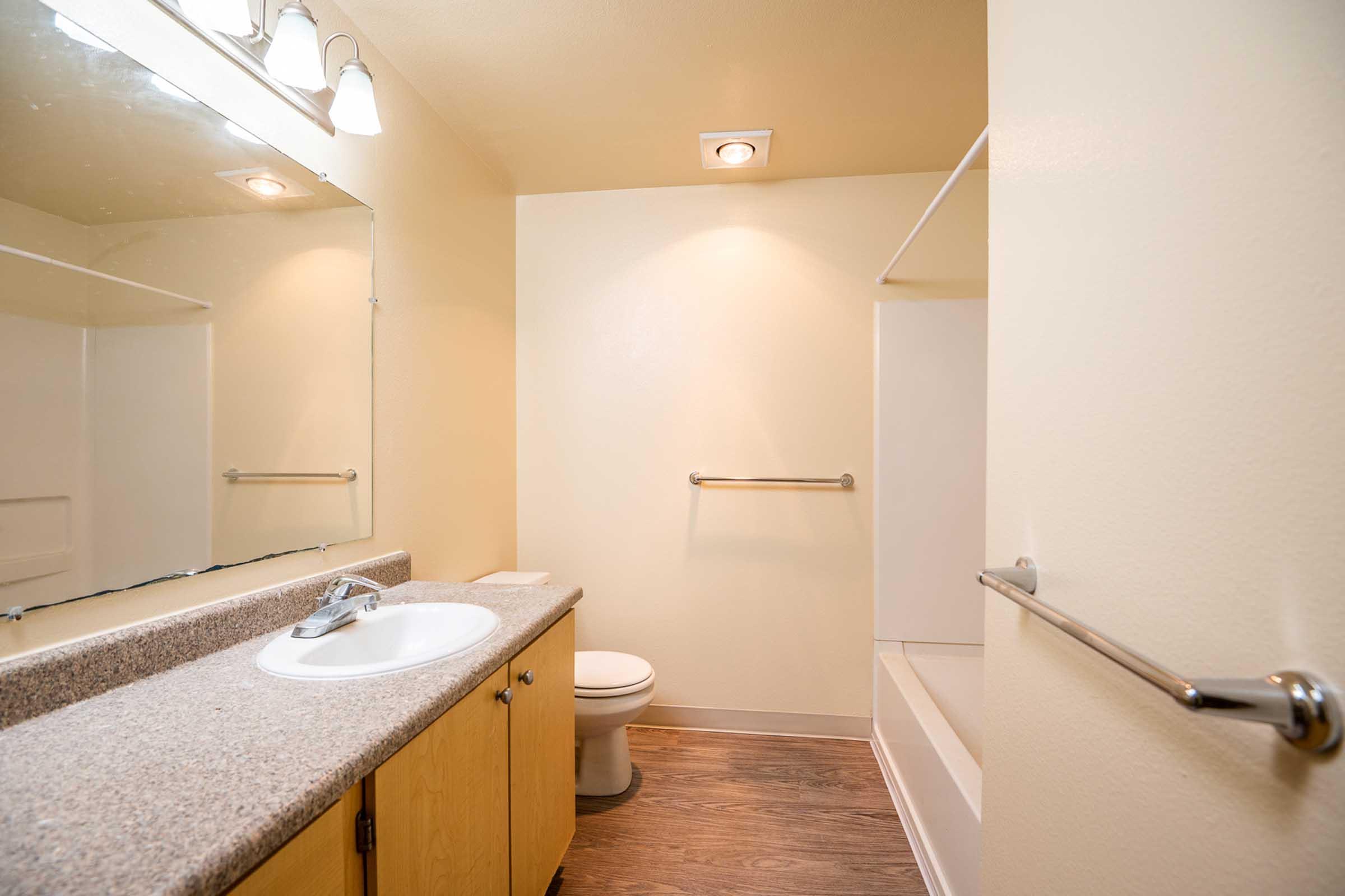 A modern bathroom featuring a compact design with a light-colored countertop, a mirror with three light fixtures above, a sink with a faucet, a toilet, and a bathtub with a shower. The walls are painted in a soft neutral tone, and the floor has a wooden finish, creating a bright and clean atmosphere.