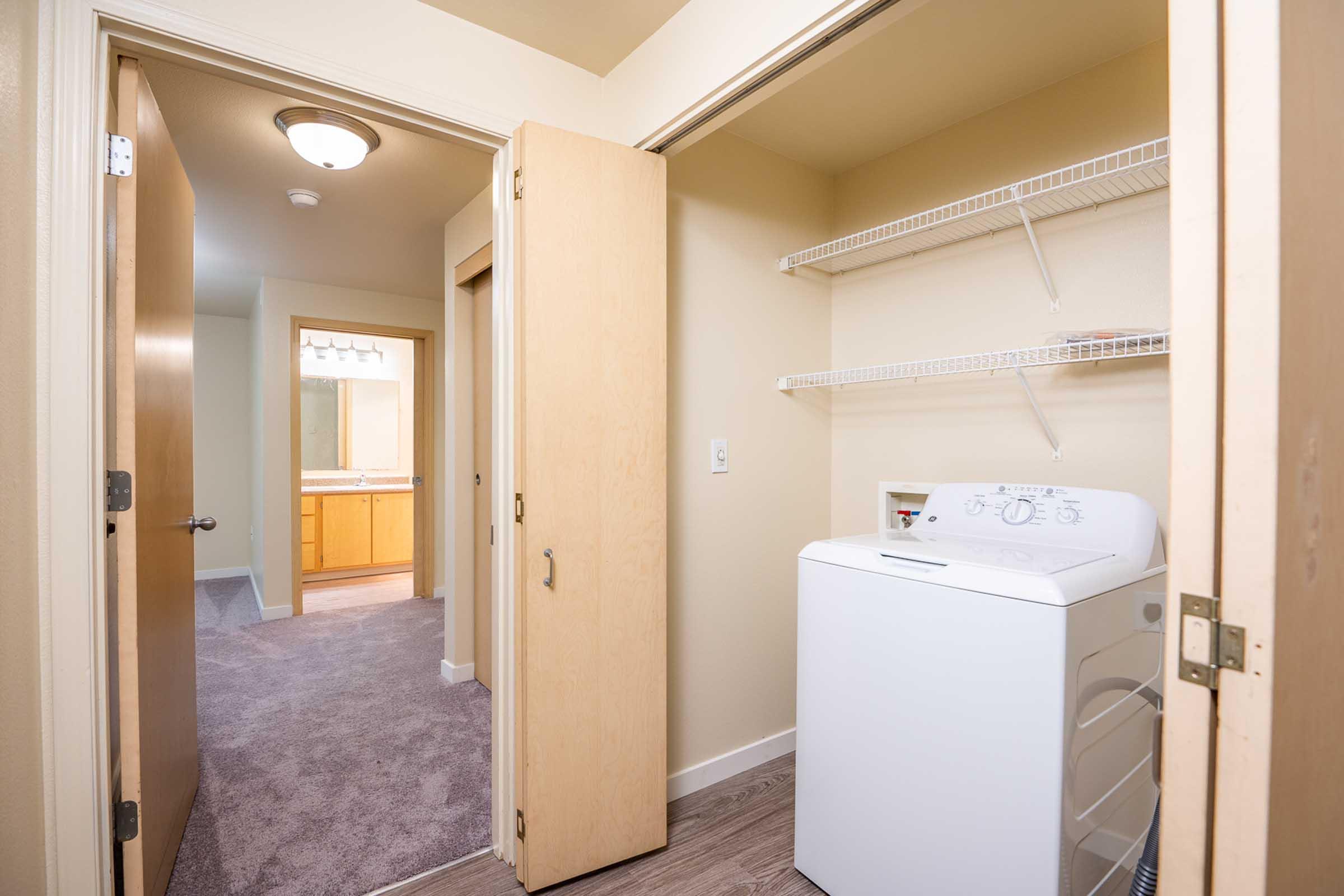 A laundry area with a white washing machine located in a hallway. Light-colored walls and carpet can be seen, along with an open door leading to a bathroom vanity area with a mirror. There is a shelf above the washing machine for storage.