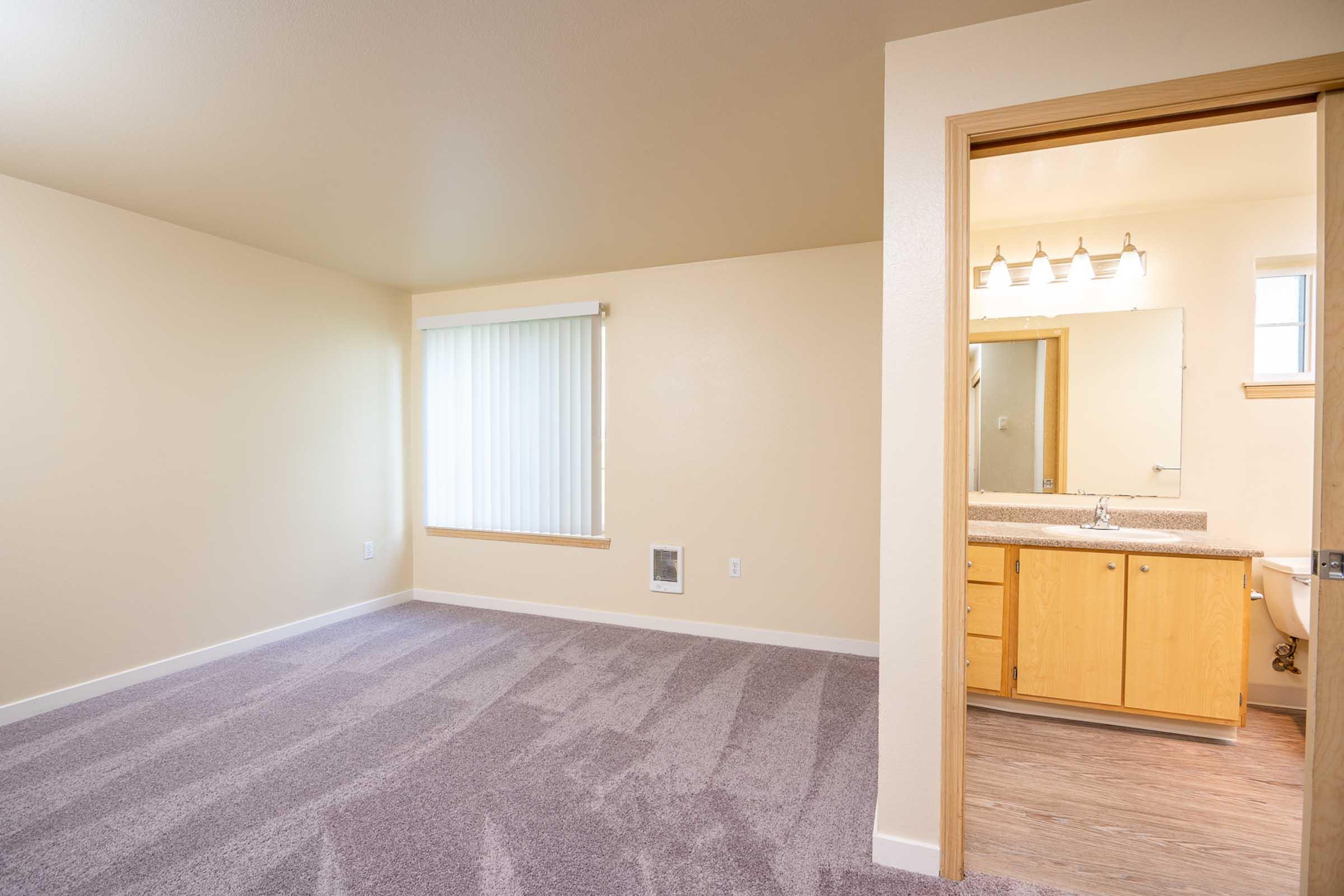 A spacious, empty room with light beige walls and soft carpet, featuring a window with vertical blinds. To the right, there's a doorway leading to a bathroom with a modern vanity and mirror, illuminated by overhead lighting. The overall atmosphere is bright and inviting.