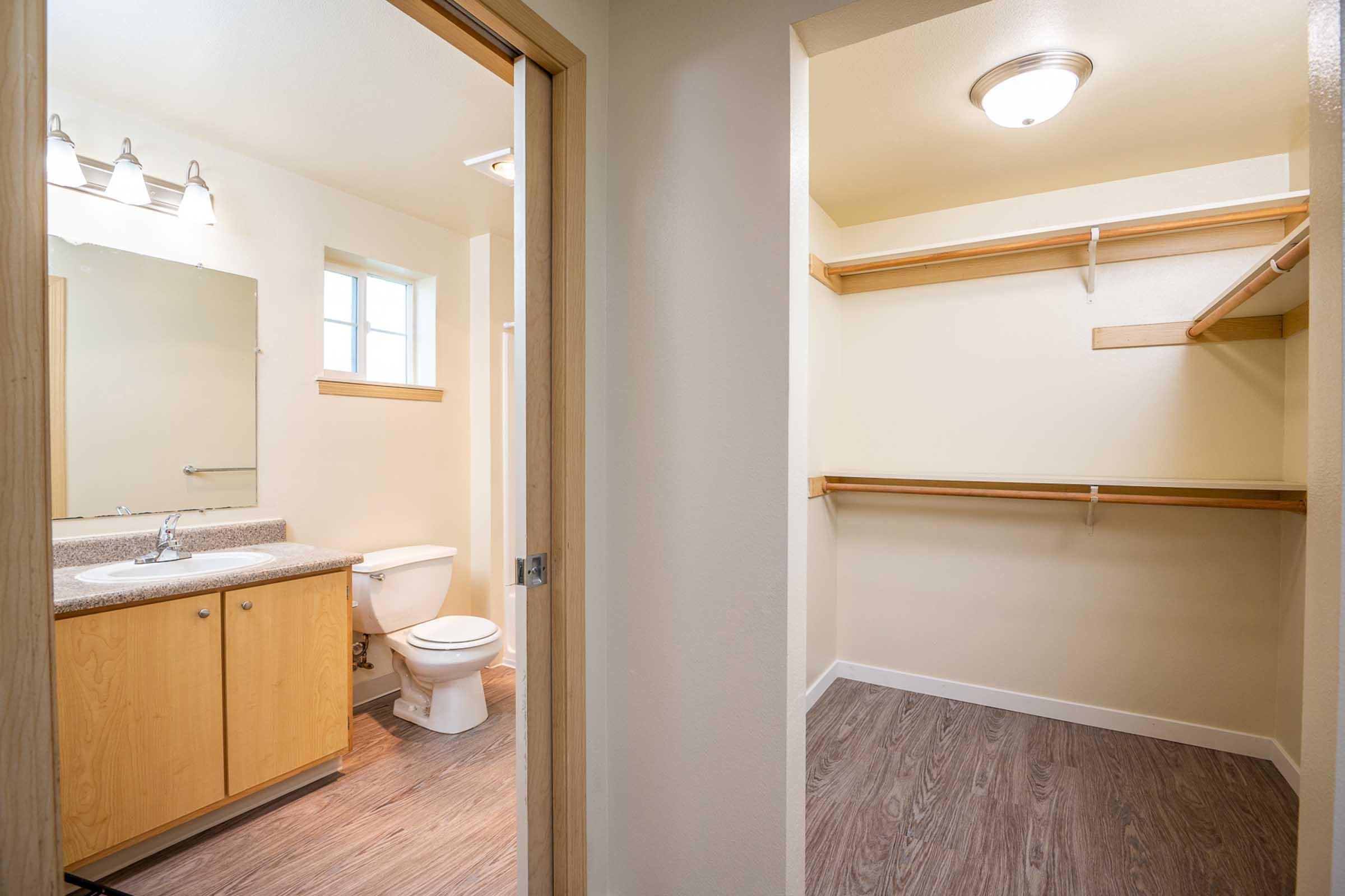 A view through an open door into a bathroom and adjoining closet. The bathroom features a sink with a mirror, a toilet, and light fixtures above. The closet has shelves and hanging space, with light-colored walls and wood flooring visible in both spaces.