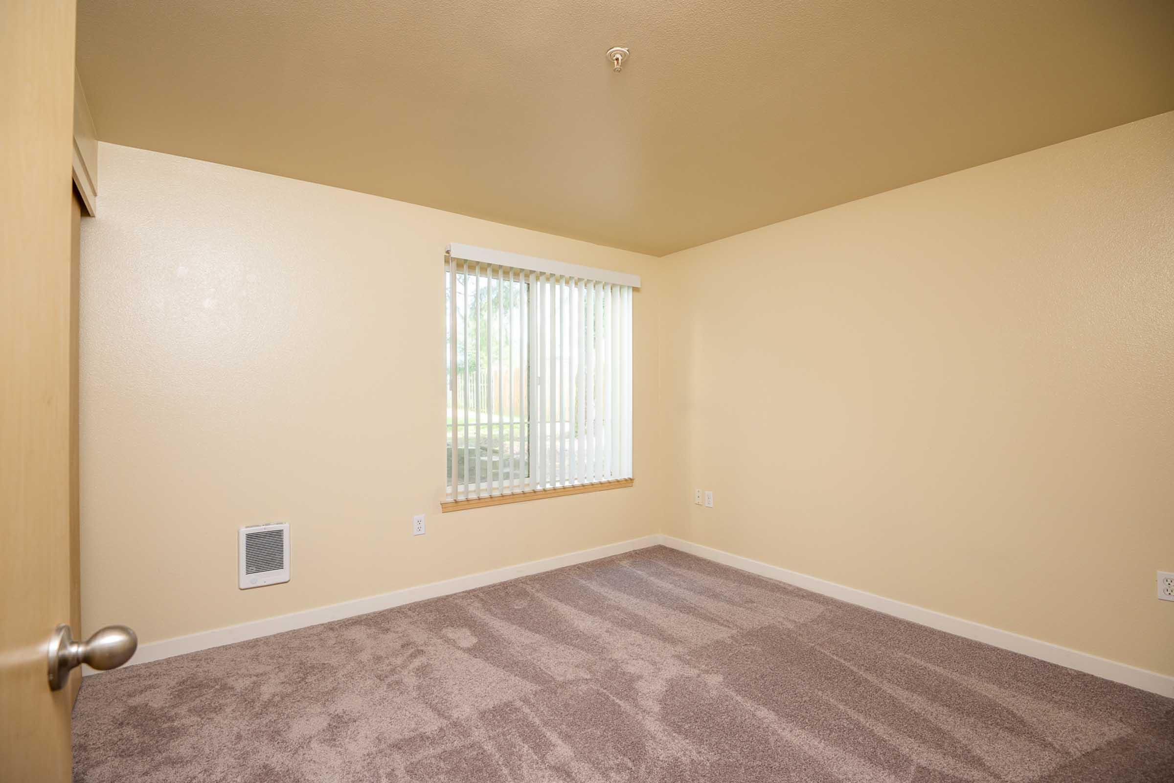Empty room with light beige walls and plush carpet. A window with vertical blinds allows natural light to enter. An air vent is visible on one wall. The room appears spacious and is ready for furnishings.