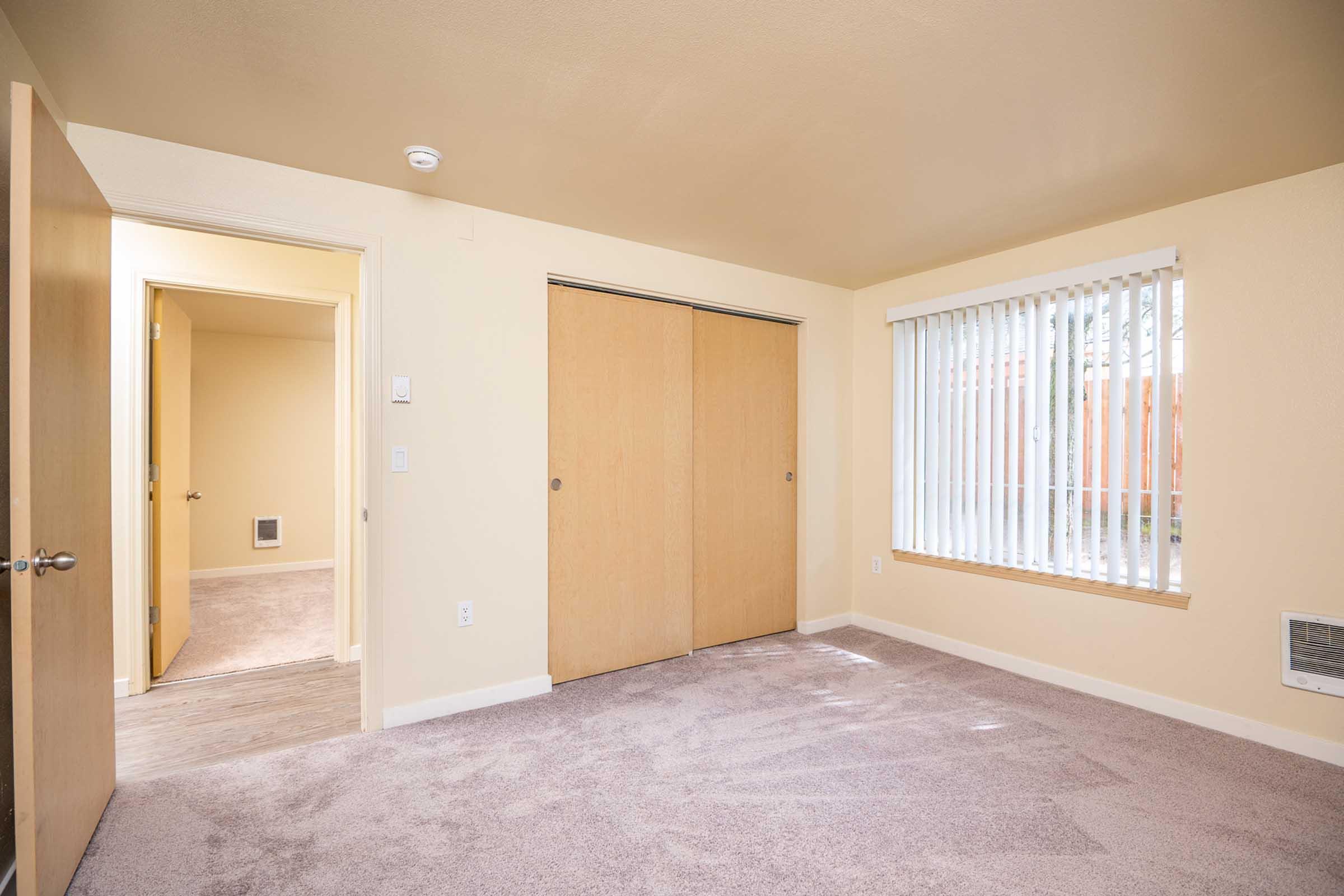 Empty room with beige walls and carpeted floor. Features a sliding closet door, a window with vertical blinds allowing natural light, and an entrance leading to another room. The door to the left opens into a small hallway, enhancing the spacious feel of the area.