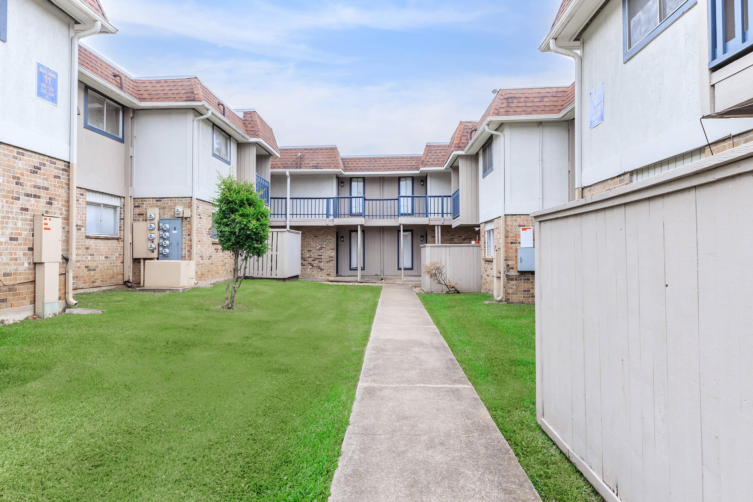 A well-maintained apartment complex featuring two-story buildings arranged around a grassy courtyard. A concrete pathway leads through the center, flanked by well-trimmed lawns and small shrubs. The buildings have light-colored exterior walls with red patterned roofs and multiple windows.