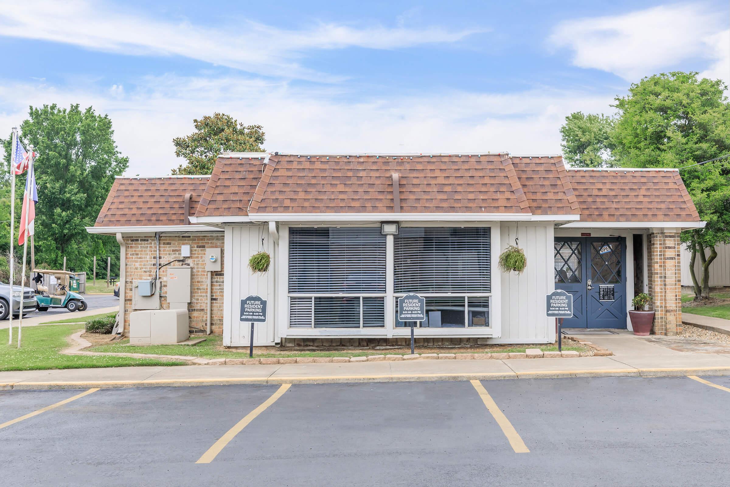 A single-story brick building with a sloped, brown tile roof, featuring large windows with blinds. The front has potted plants and several signs on either side. In the background, there are trees and a clear blue sky. The parking lot in front has marked spaces.