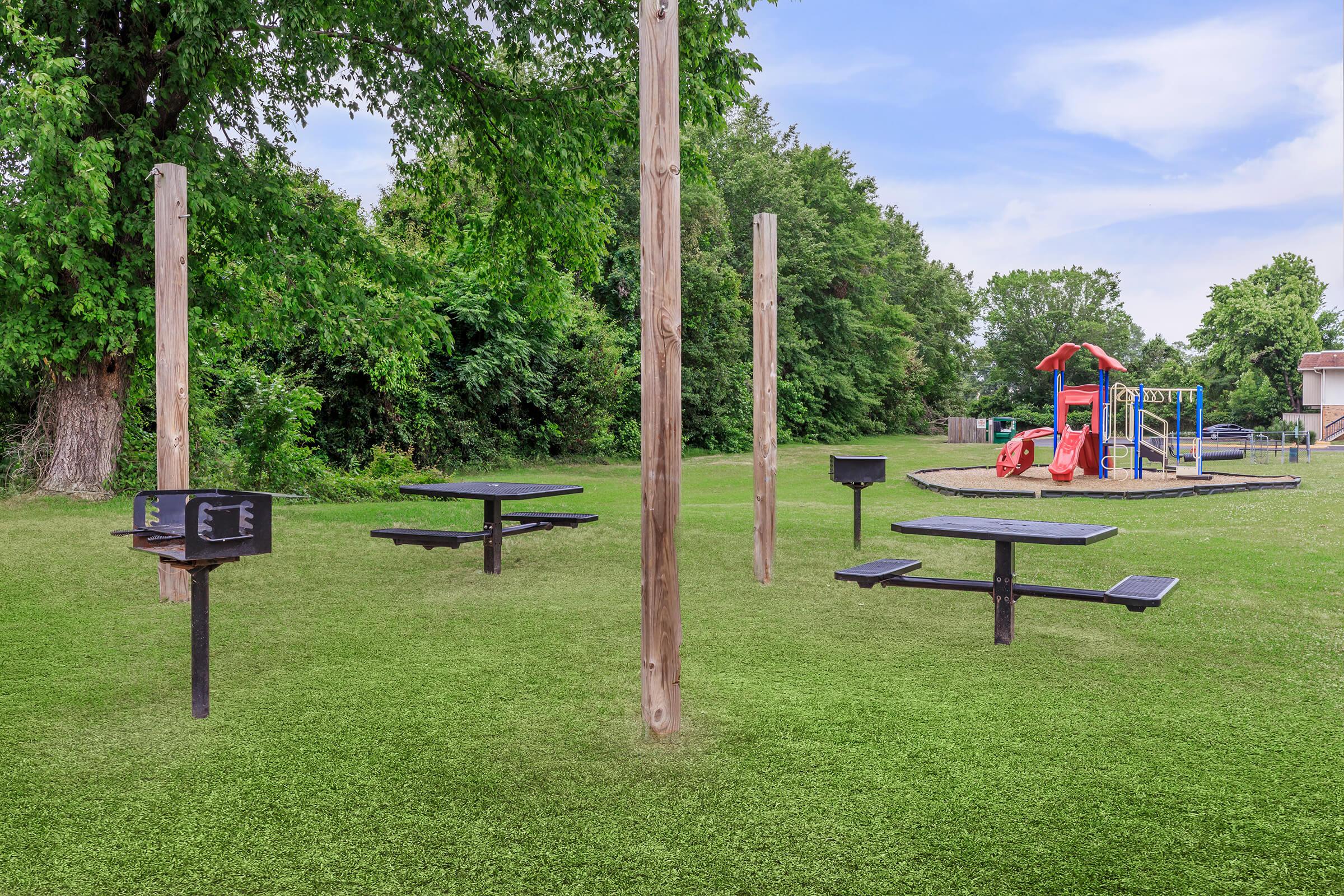 A grassy park area featuring several picnic tables and grills, with a playground in the background. The playground includes a red slide and climbing frame, surrounded by tall trees and open green space.