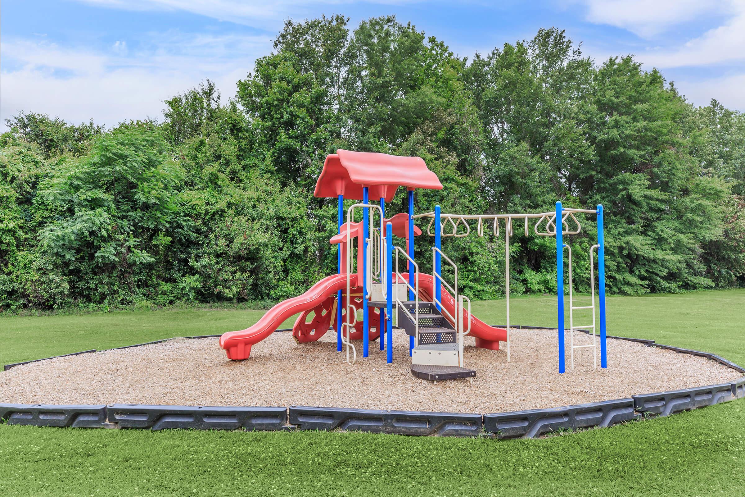 A colorful playground featuring a red slide, climbing structures, and swings, situated on a soft, sandy surface. Lush green trees create a natural backdrop, making it a cheerful outdoor space for children to play and explore.
